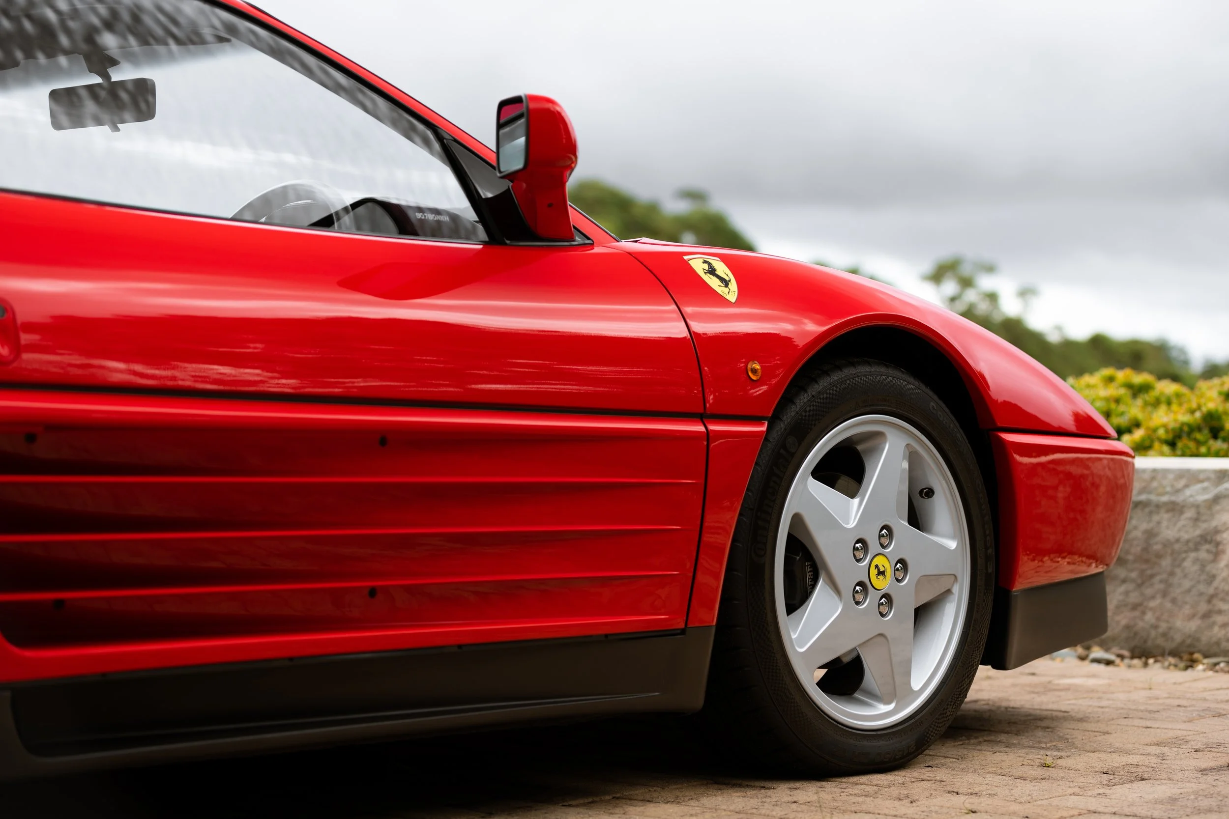 Close-up of a red Ferrari sports car, showing part of the front wheel, side mirror, and Ferrari logo on the fender, parked outdoors.