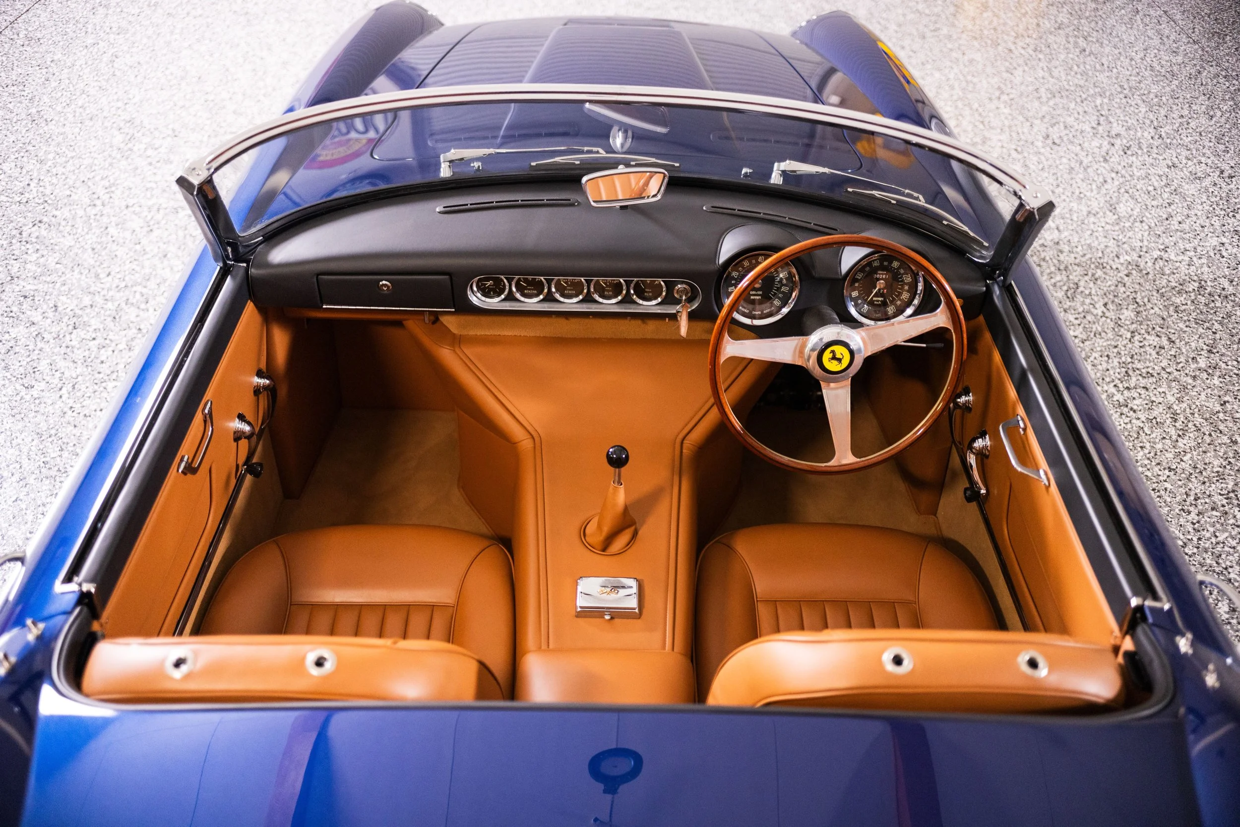 Interior view from above of a vintage blue sports car with tan leather seats, a wood steering wheel, and a dashboard with multiple gauges.