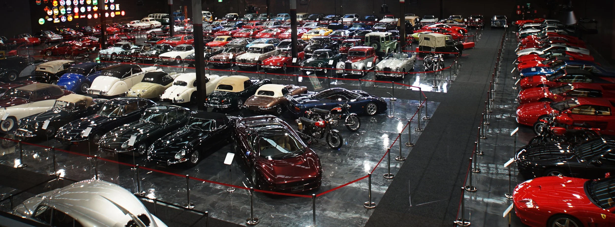 A large indoor classic car museum or showroom with rows of vintage and sports cars, including convertibles, sedans, and motorcycles, all on display behind red ropes with signage, with a collection of automotive memorabilia on the wall.