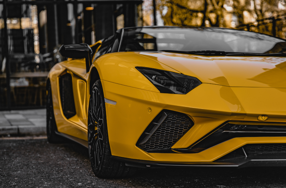 A yellow luxury sports car parked outdoors, with a dark background and autumn trees reflected in its surface.