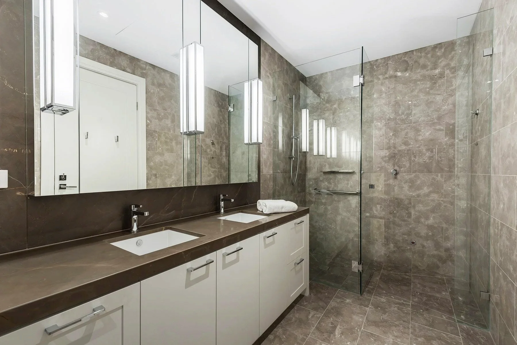 A private apartment's bathroom with large mirror, dual sinks, stone countertop, wall to ceiling tiles, walk-in shower with glass door, and wall-mounted lighting fixtures.