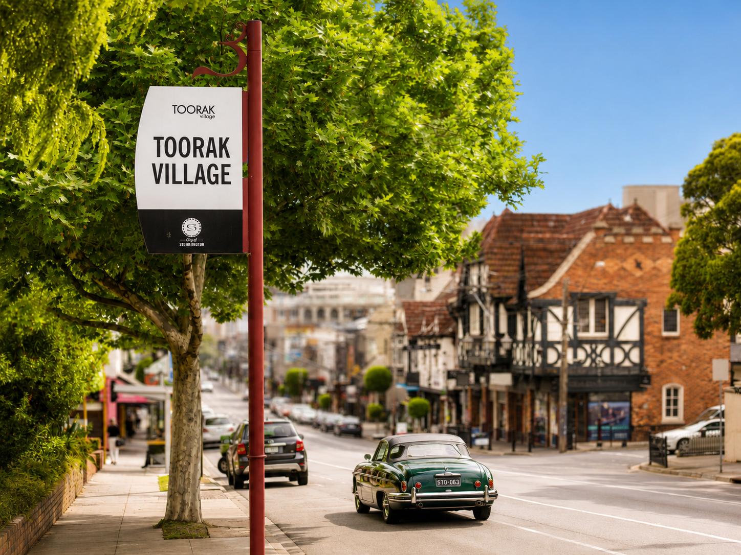 Street view of Toorak Village with a vintage green car and traditional shops, street lined with trees and a clear blue sky.