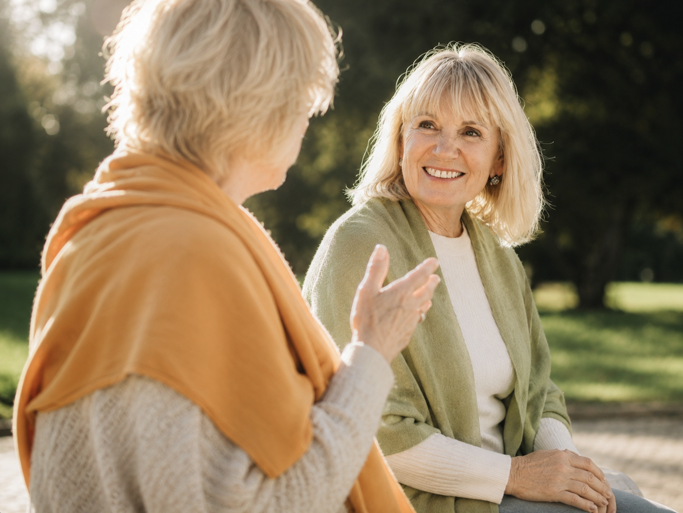 Two older women sitting outdoors in a park, engaged in conversation with sunny weather and green trees in the background.