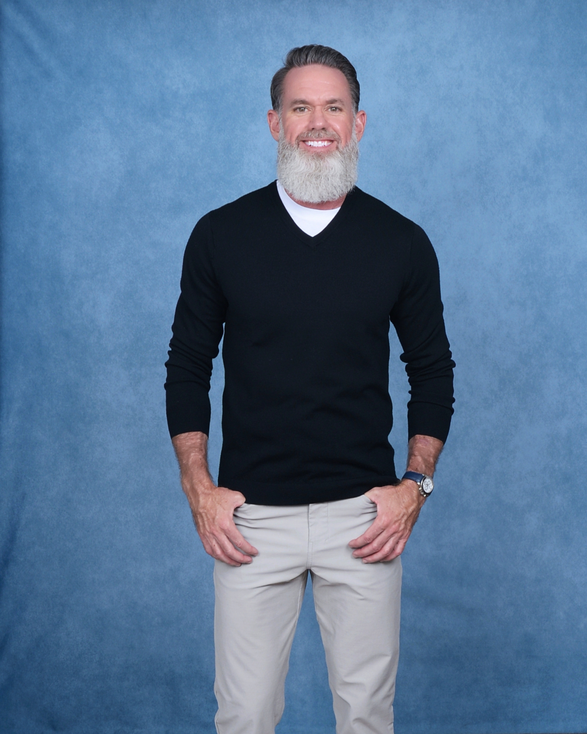Portrait of founder Matthew Thrasher, wearing a black V-neck sweater over a white shirt and beige pants, standing against a solid blue background.