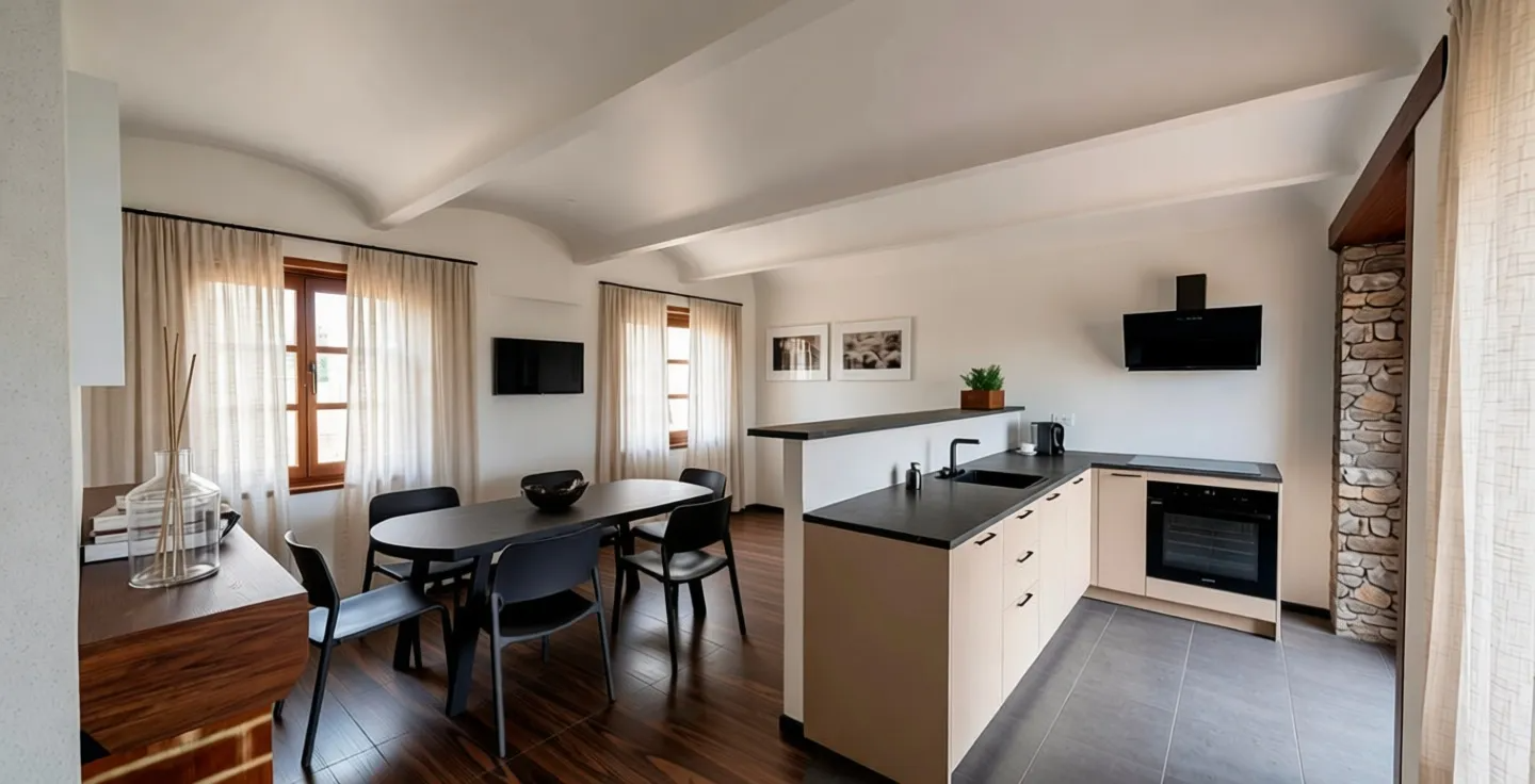 Interno di una cucina e sala da pranzo con pareti bianche, finestre con tende chiare, tavolo nero con sedie, cucina con superfici nere e ante chiare, parete con effetti di pietra per un camino, televisori sulla parete, decorazioni e piante.