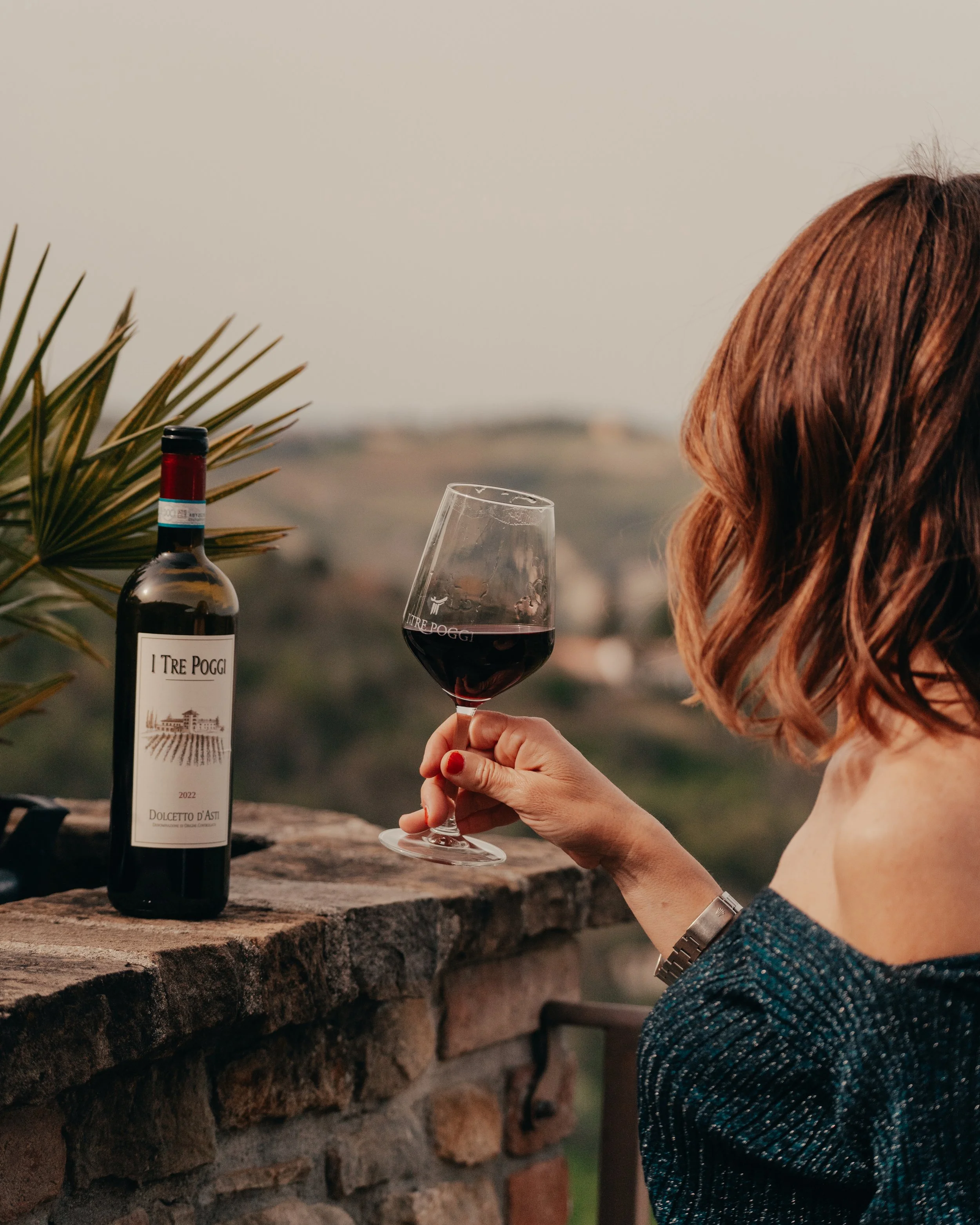 Una donna con i capelli castani che tiene un bicchiere di vino rosso, in un balcone di pietra, con una bottiglia di vino sul parapetto e un paesaggio collinare sullo sfondo