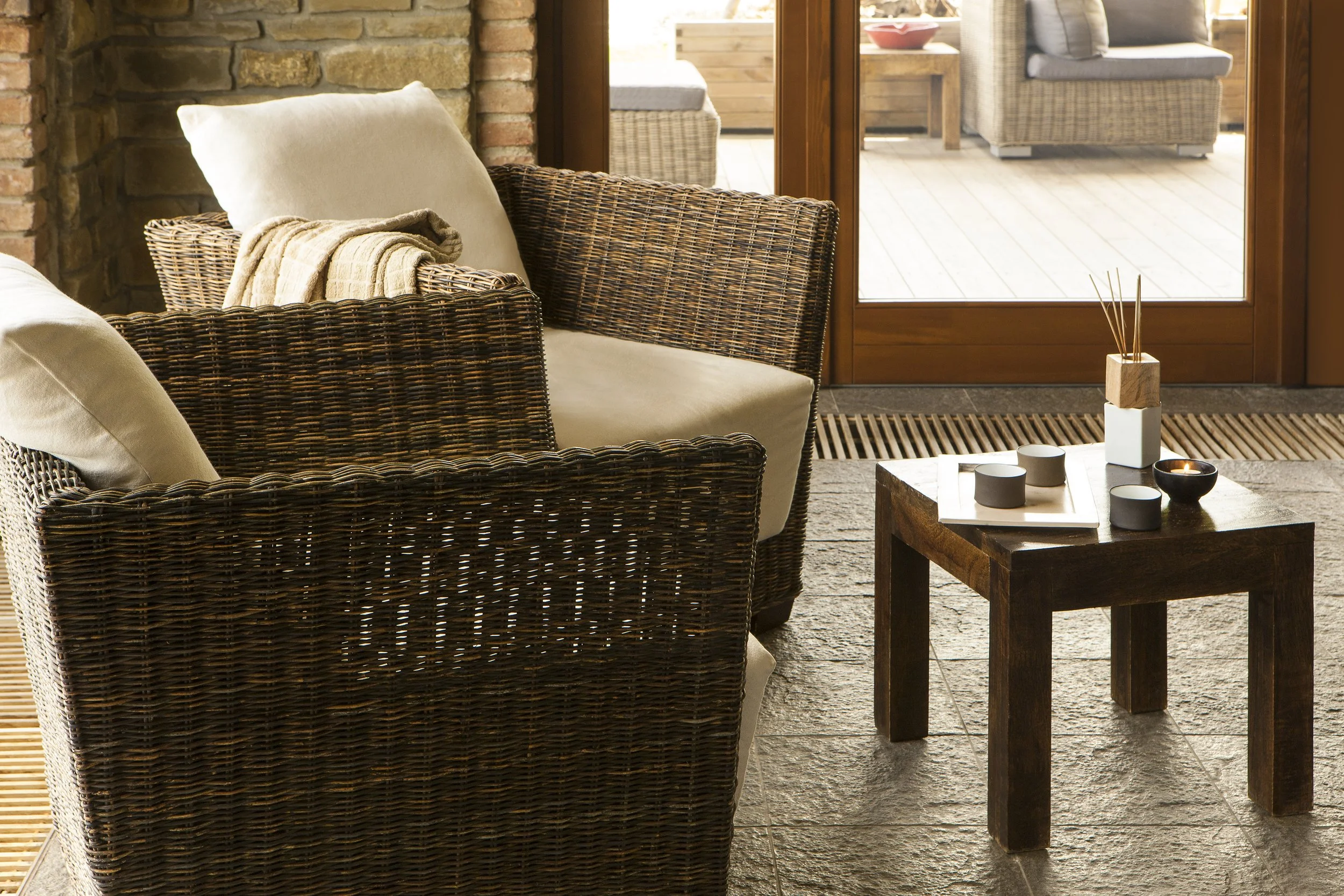 Soggiorno con sedie in vimini, cuscini beige, tavolino in legno con candele, porta finestra che dà su un balcone con sedie e tavolino in vimini