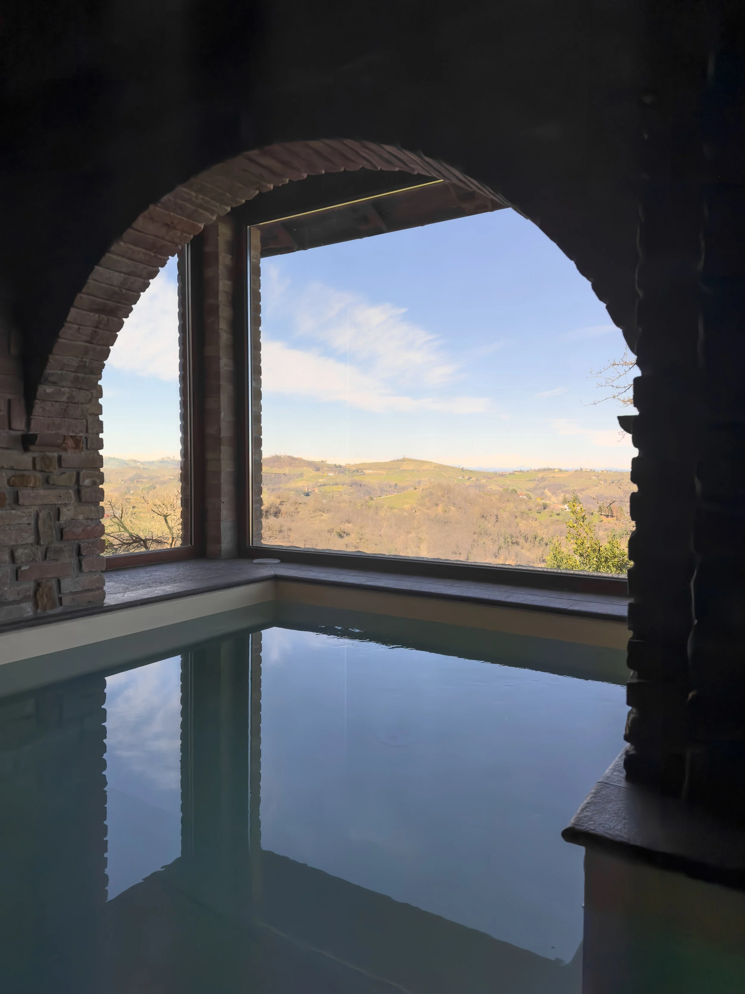 Vista di una finestra con un paesaggio collinare e cielo azzurro, riflesso in una vasca da bagno, con un muro di mattoni rustico all'interno dell'ambiente.