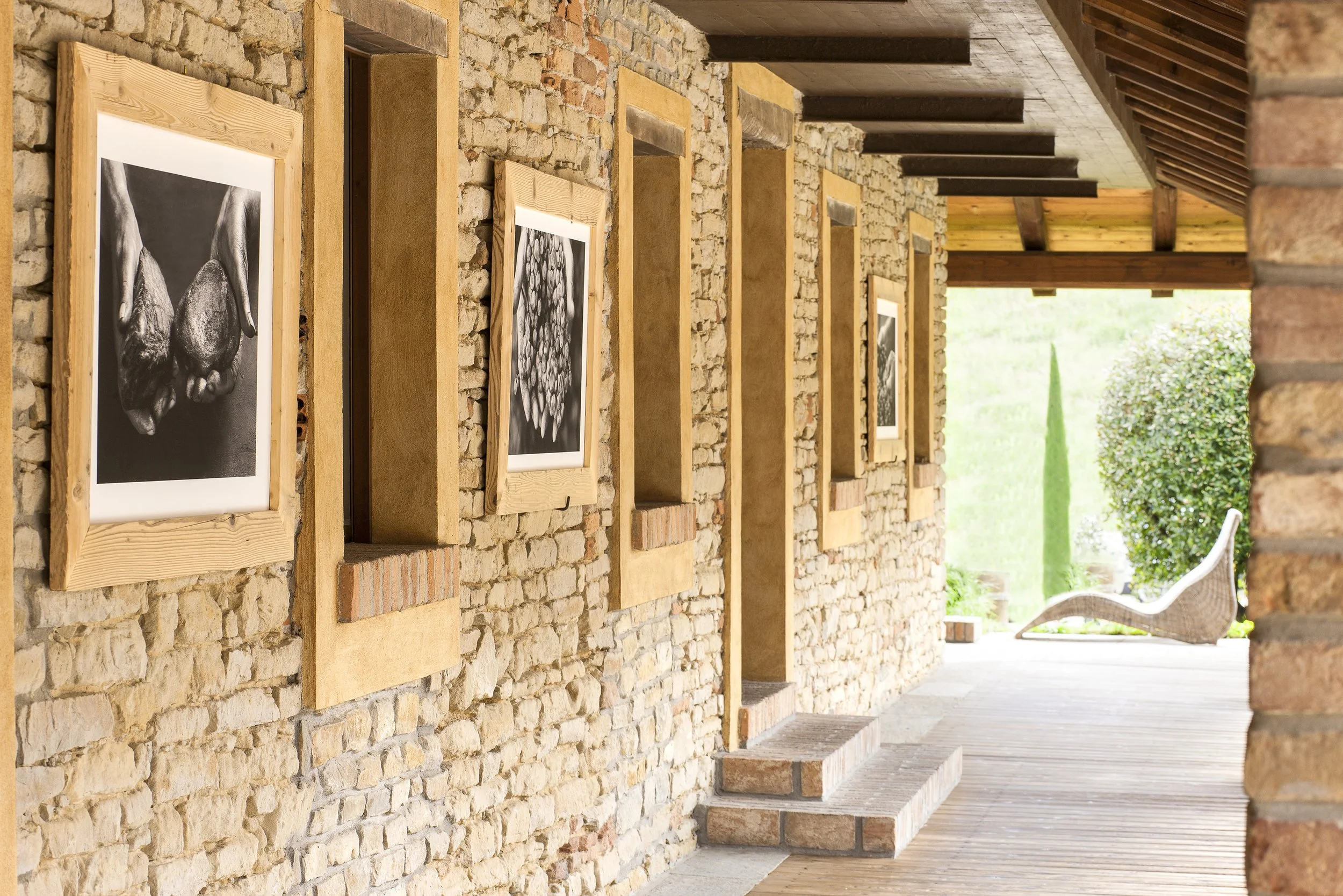 Una parete di mattoni con tele extensive di fotografie in cornici di legno, un'area veranda con sedie e fiori, e un paesaggio aperto con cespugli e alberi.