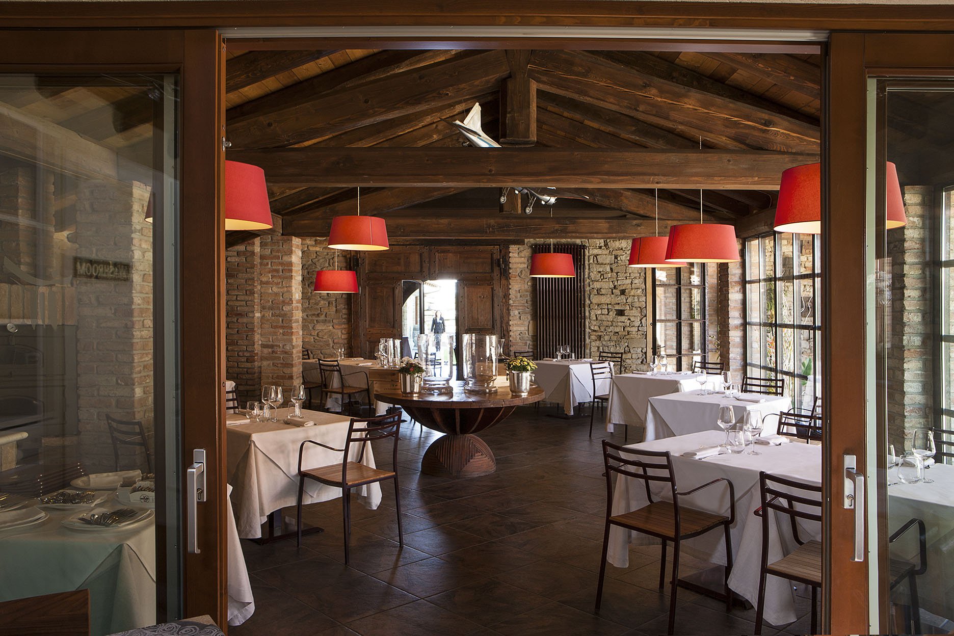 Una sala da pranzo elegante con tavoli coperti da tovaglie bianche, bicchieri e posate, illuminata da lampade rosse a soffitto, pareti in pietra e grandi finestre che lasciano entrare luce naturale.