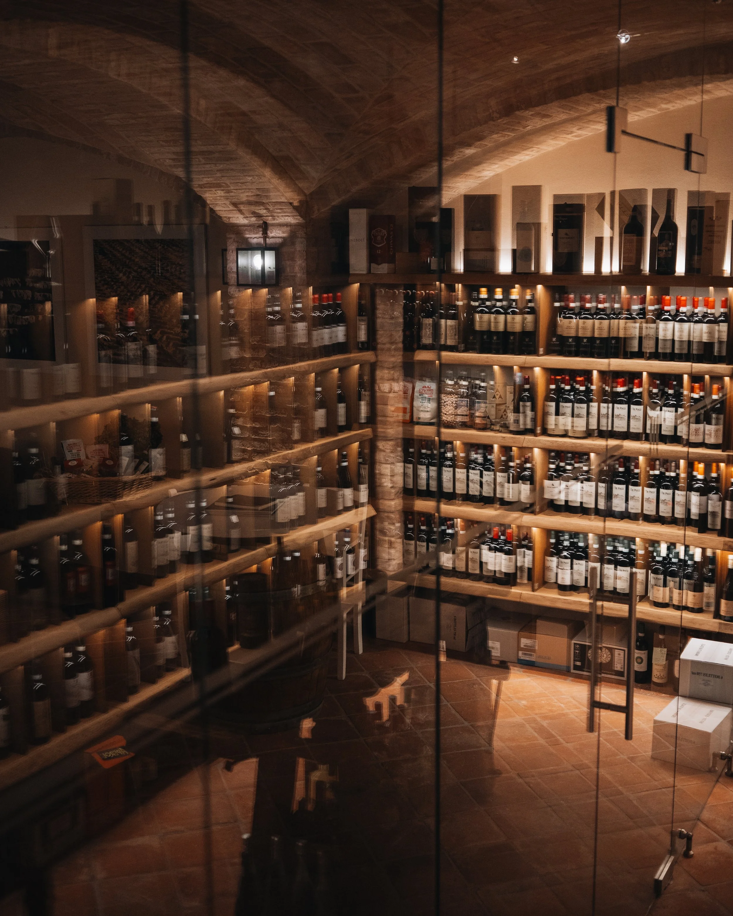Interno di una enoteca o negozio di vini con scaffali pieni di bottiglie di vino in legno, luci calde e un ambiente rustico con soffitto in pietra e pavimento in ceramica.