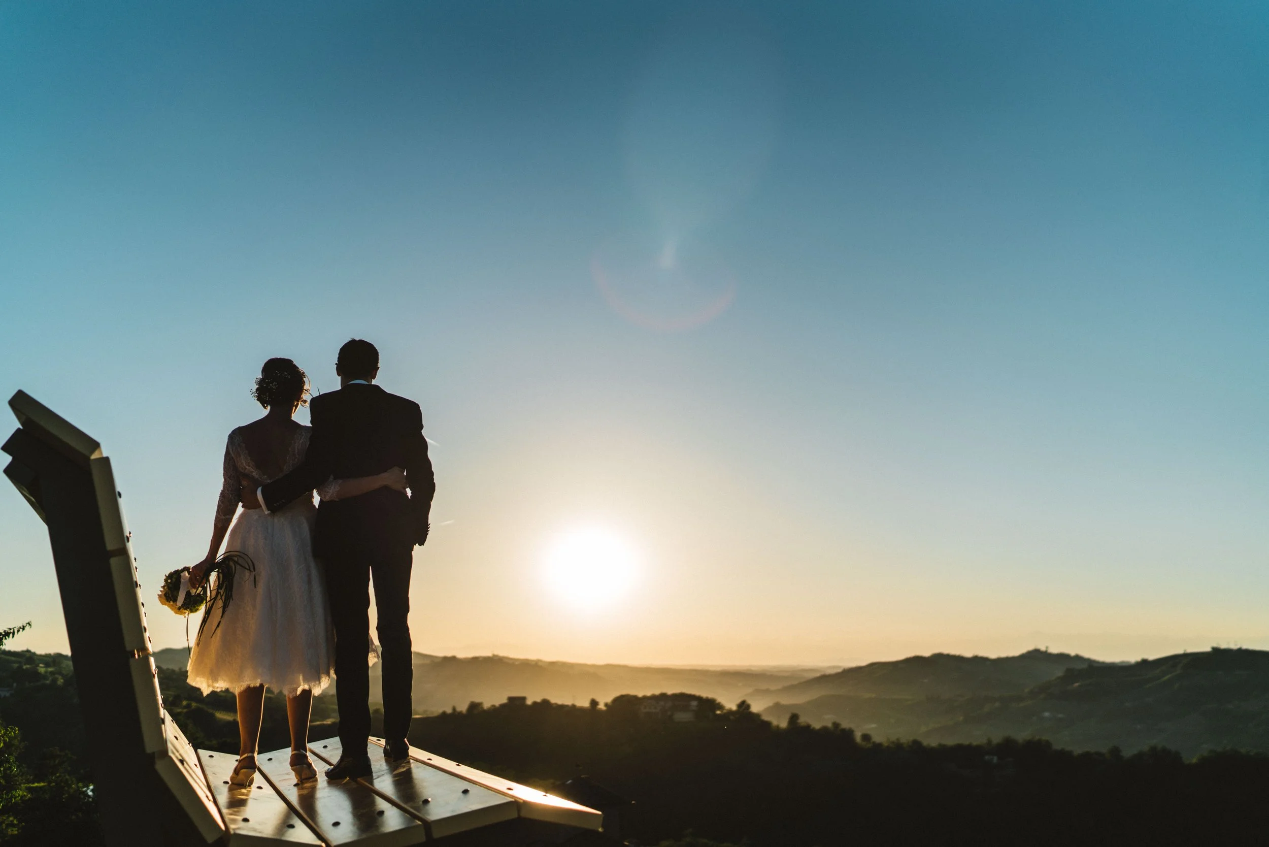 Una coppia di sposi si fa fotografare al tramonto su una terrazza panoramica, con il paesaggio collinare sullo sfondo.