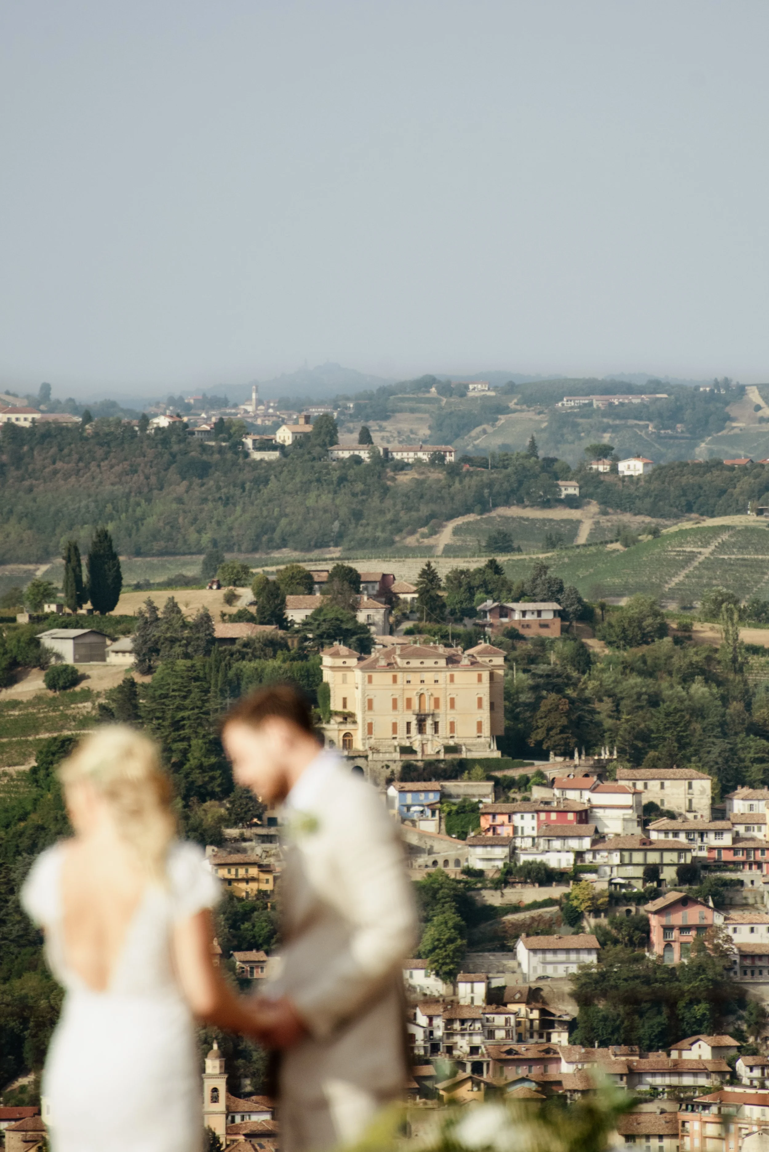 Vista panoramica di un paesaggio collinare con case e alberi, alcune persone in primo piano sono sfocate.