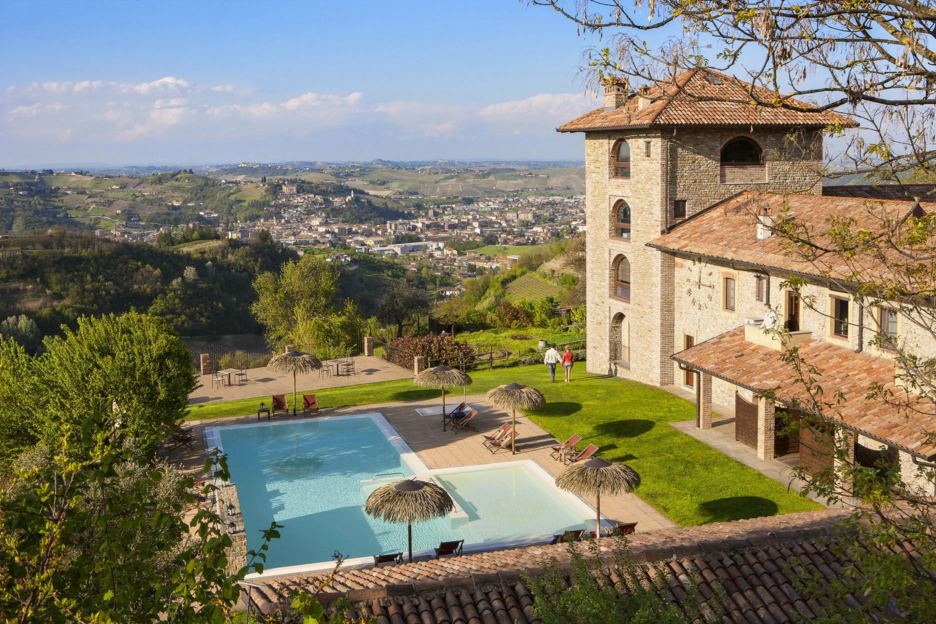 Scorcio di una casa in stile rustico con torretta e tetto in terracotta, piscina all'aperto con ombrelloni di paglia, giardino verde con alberi, veduta panoramica di una città e colline in lontananza.