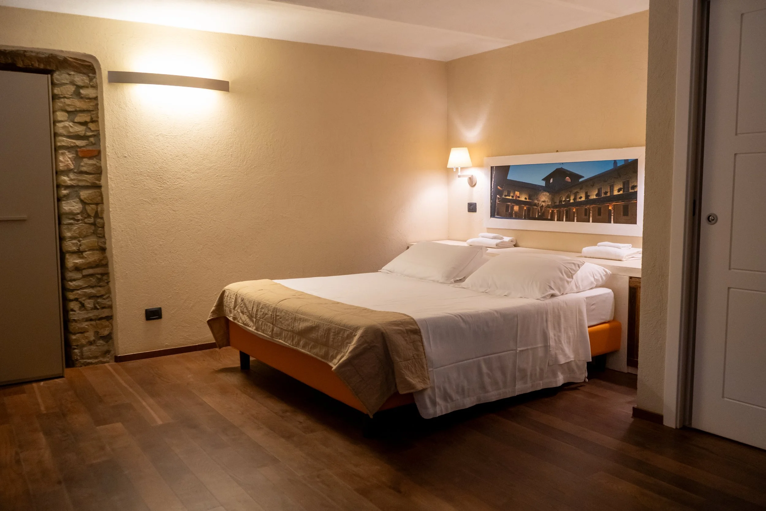 Camera da letto con letto matrimoniale, pareti beige, lampada da parete accesa, quadro sopra il letto che mostra un edificio storico di notte, pavimento in legno.