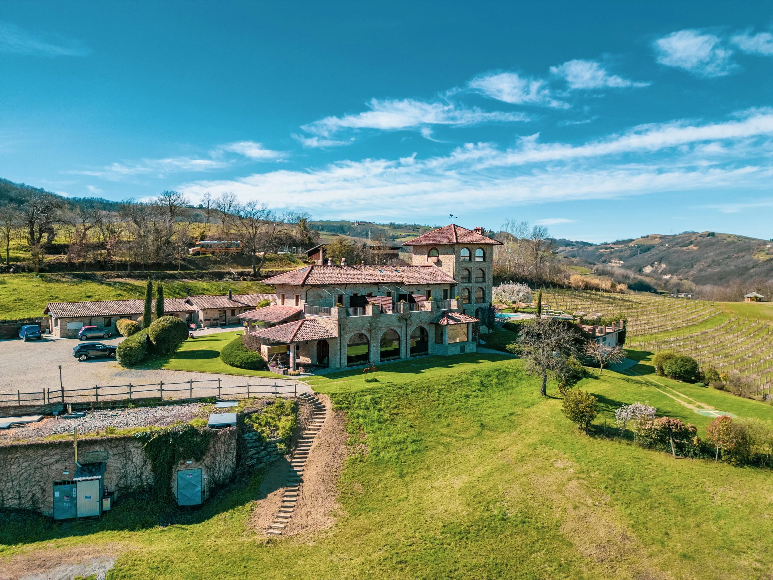 Vista panoramica di una casa in stile rustico con tetto a tegole rosse, situata in mezzo ad un paesaggio collinare verde con alberi, vigneti e un cavallo sulla prato. Cielo azzurro con nuvole sparse.