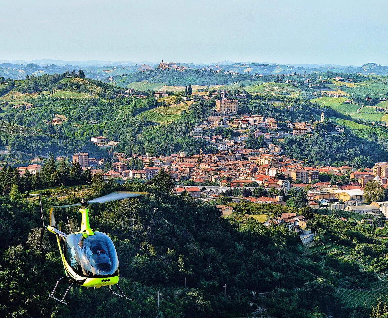 Un paesaggio collinare con un villaggio medievale, molte case e chiese, circondato da vigne e verdi colline, vista dall'alto con un elicottero giallo e nero in primo piano
