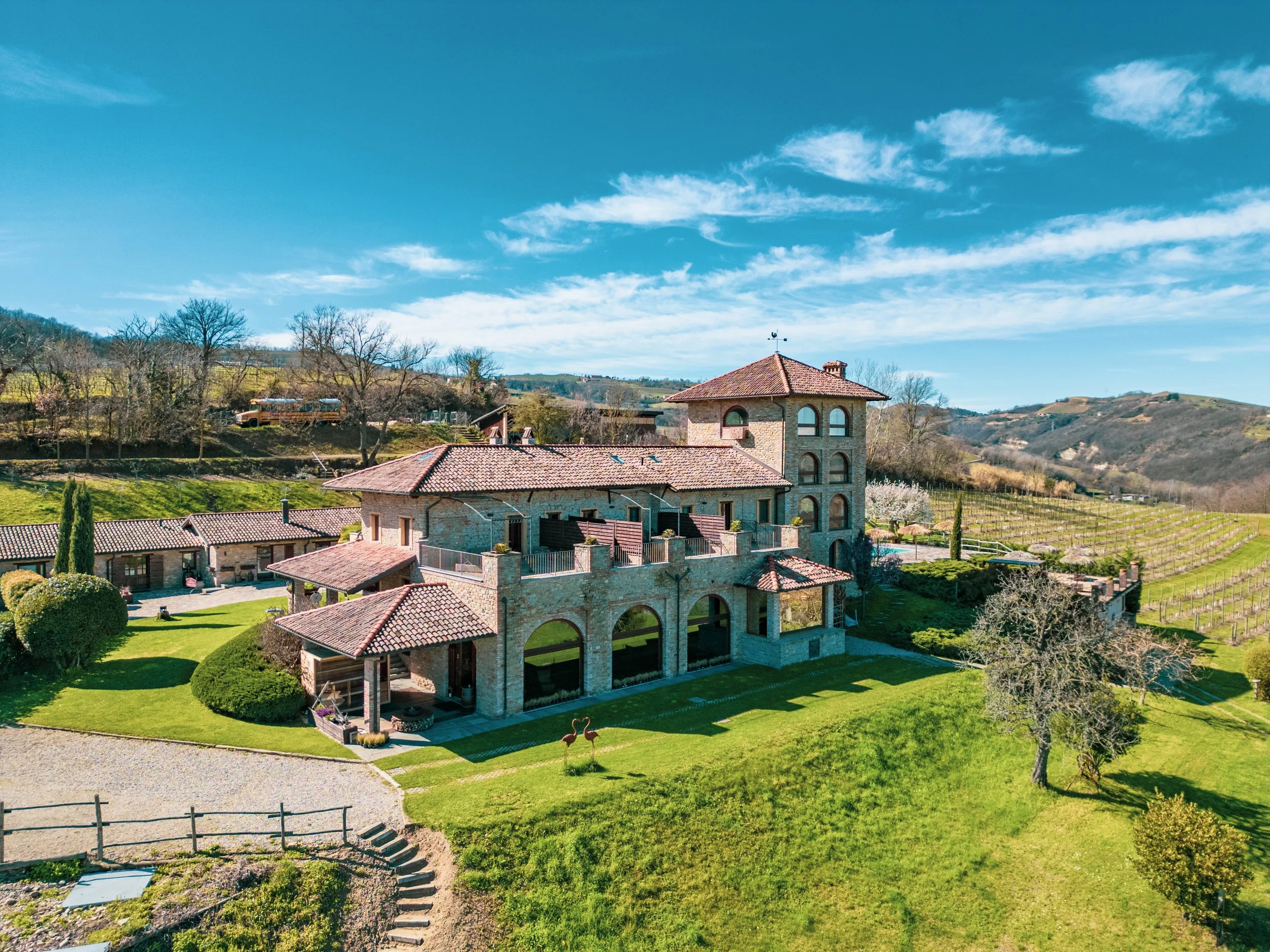 Paesaggio rurale con case in pietra, giardini curati, alberi, colline e strade di campagna sotto un cielo azzurro con alcune nuvole.