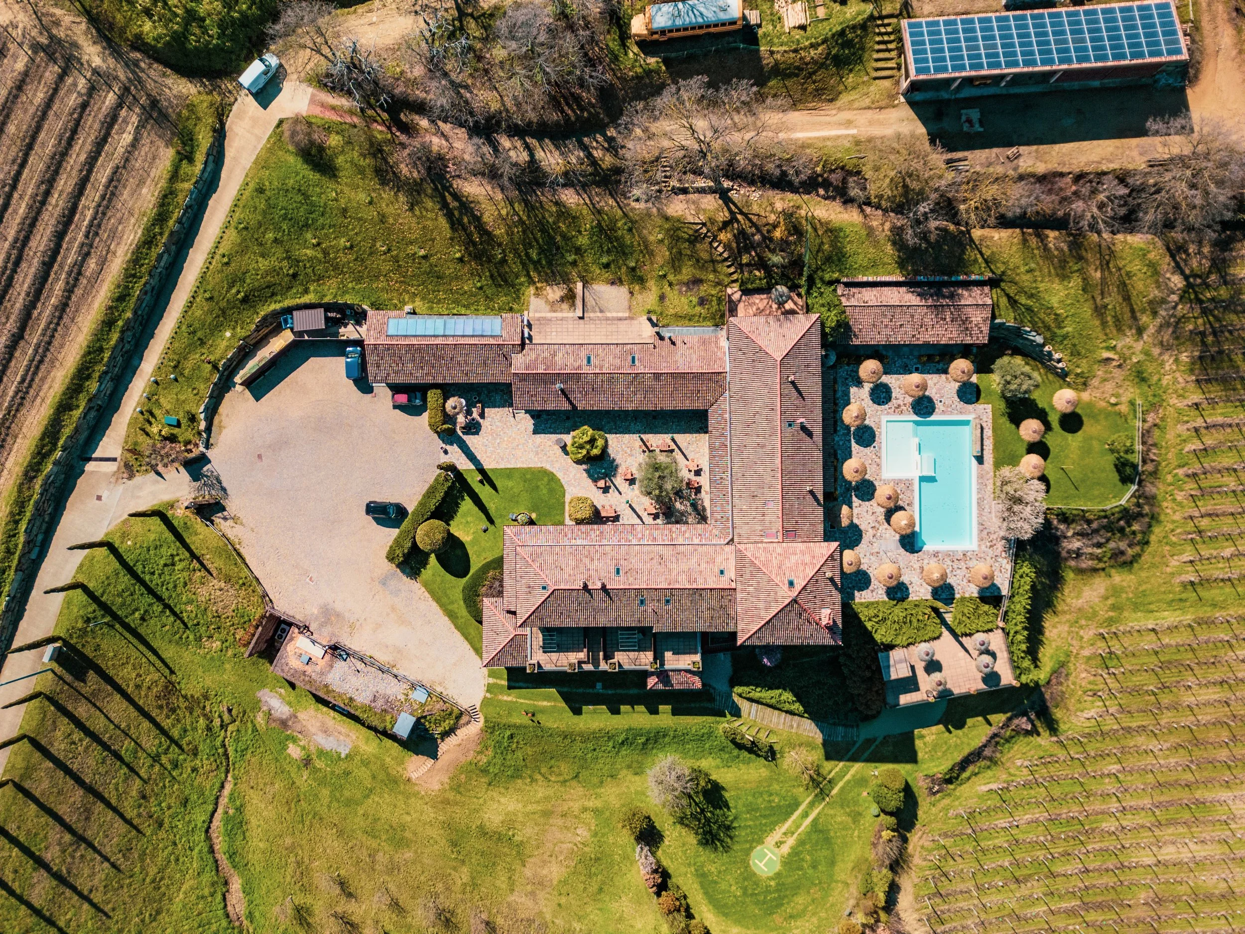Veduta aerea di una proprietà con case, piscina e giardino, circondata da verde e alberi, con un'area di parcheggio e strada.