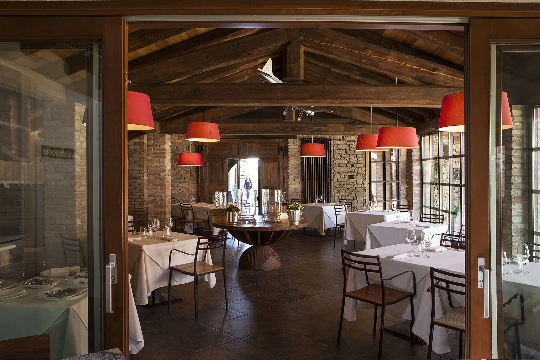 Interno di un ristorante rustico con tavoli apparecchiati, illuminazione con lampade rosse, pareti di mattoni e soffitto in legno, finestrature grandi.