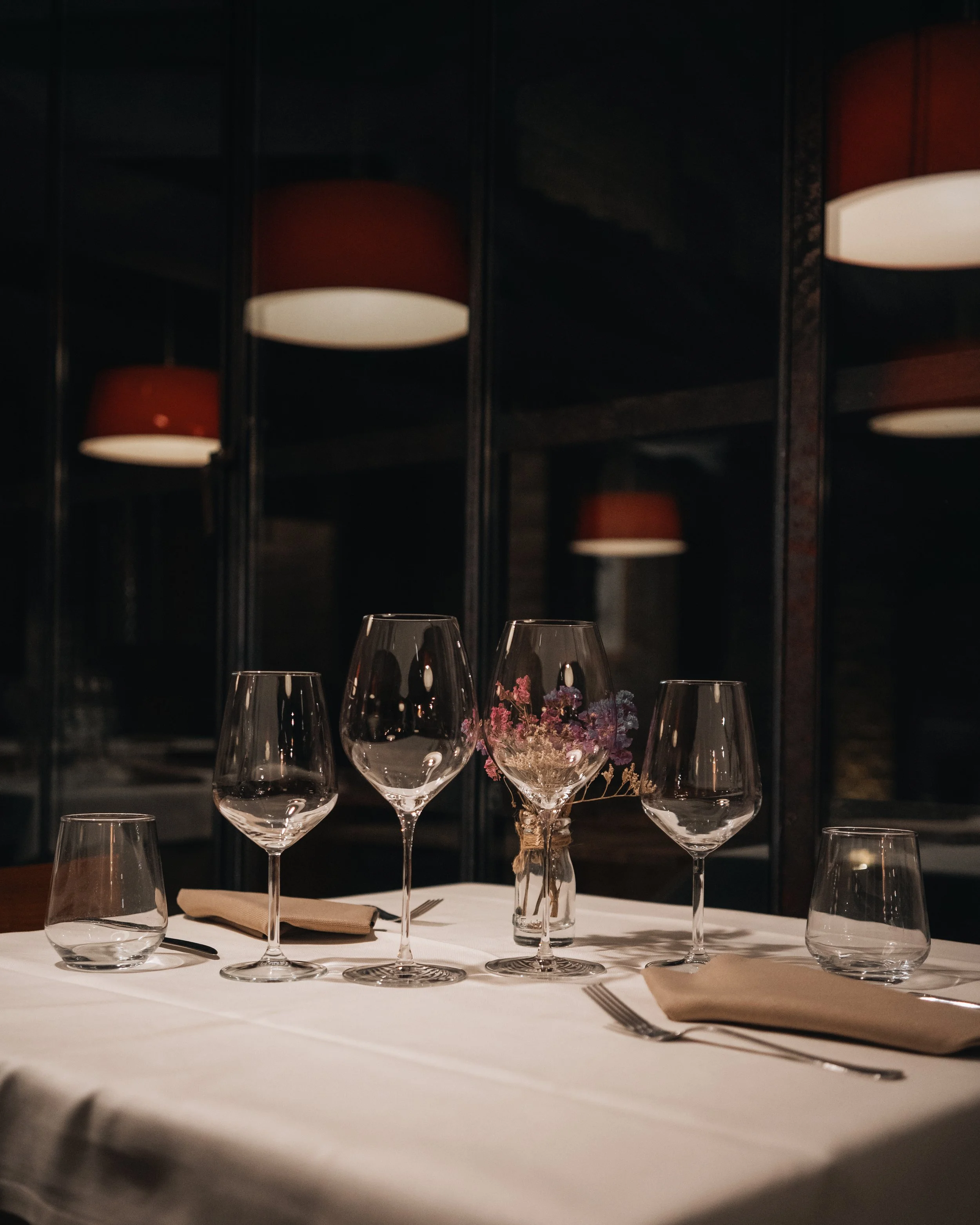 Tavola apparecchiata con sei bicchieri di vino vuoti, una piccola ciotola di fiori e tovaglioli abbinati, in un ambiente elegante con luci soffuse e lampade stilizzate sullo sfondo.