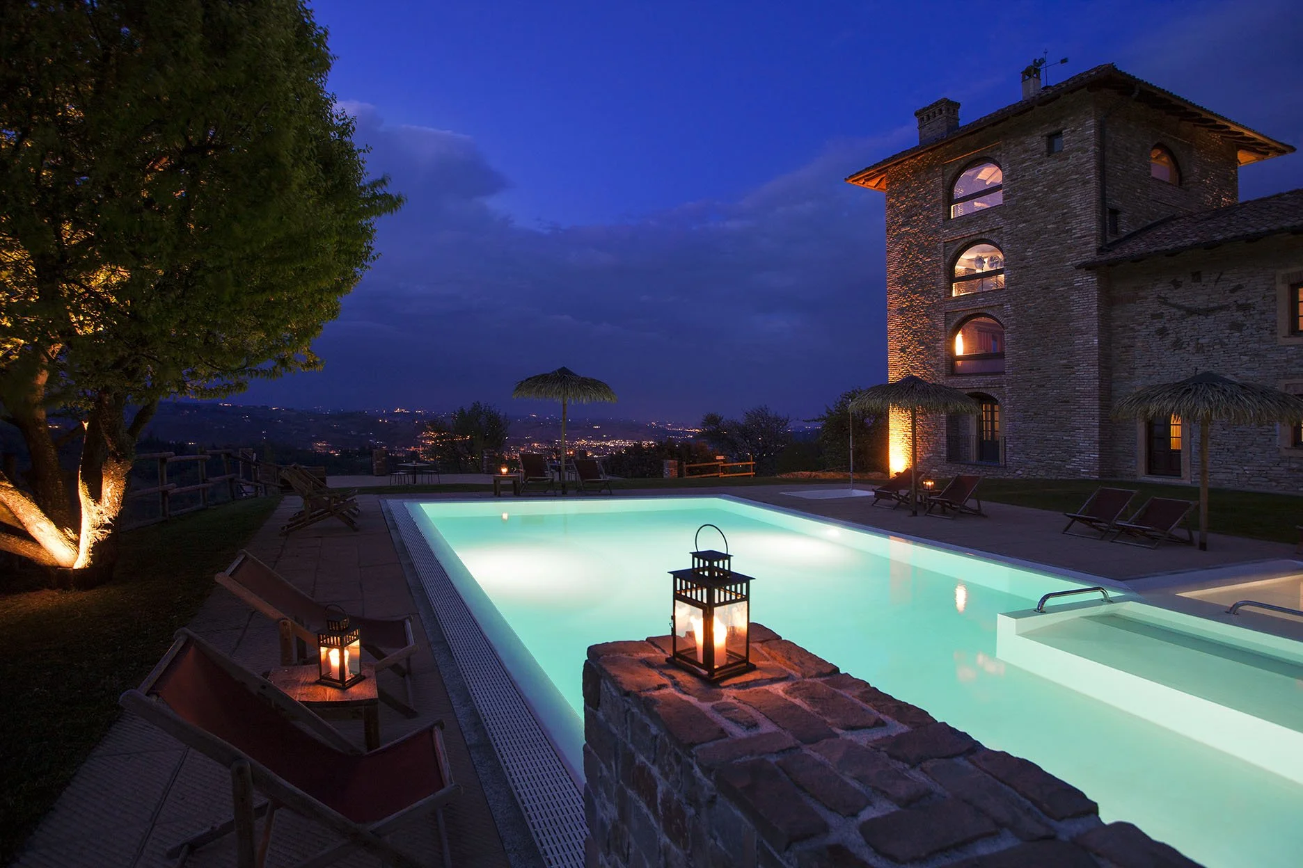 Piscina illuminata di notte con sedie a sdraio e ombrelloni di paglia, un grande edificio in pietra sullo sfondo e un cielo sereno con nuvole.