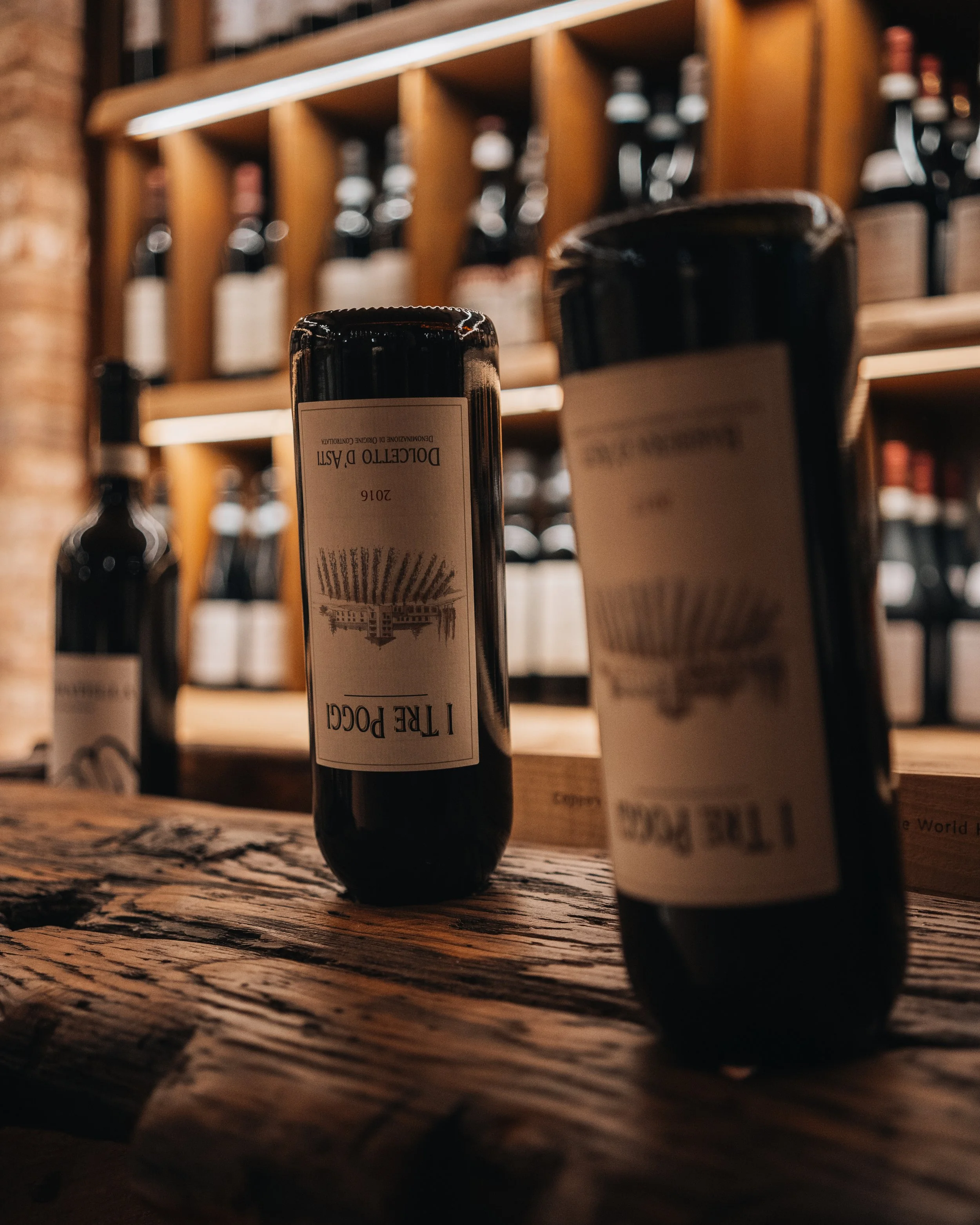 Bottiglie di vino posizionate su uno scaffale di legno in una enoteca o ristorante, con un'illuminazione calda e un'ambientazione accogliente.