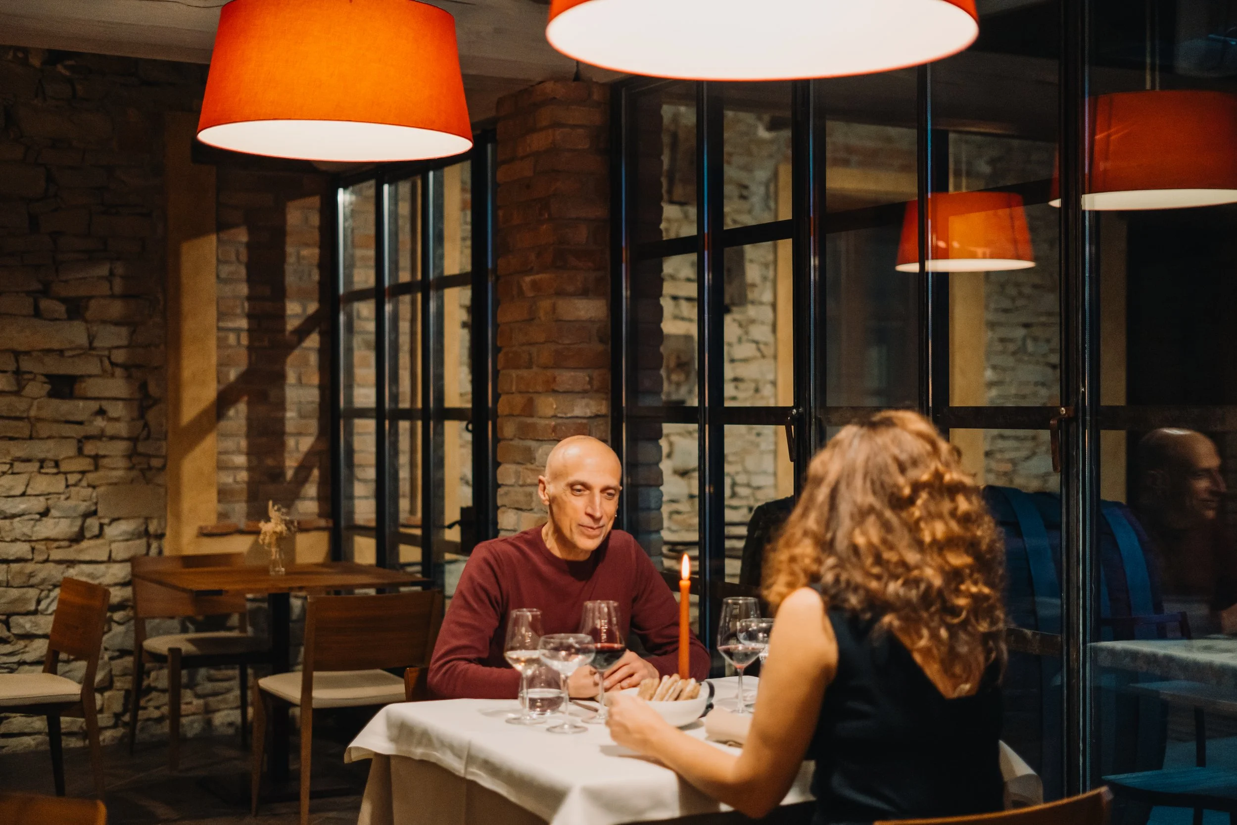 Una coppia di persone sedute a un tavolo in un ristorante romantico, con luci calde e pareti in pietra, mentre conversano e godono di una cena.