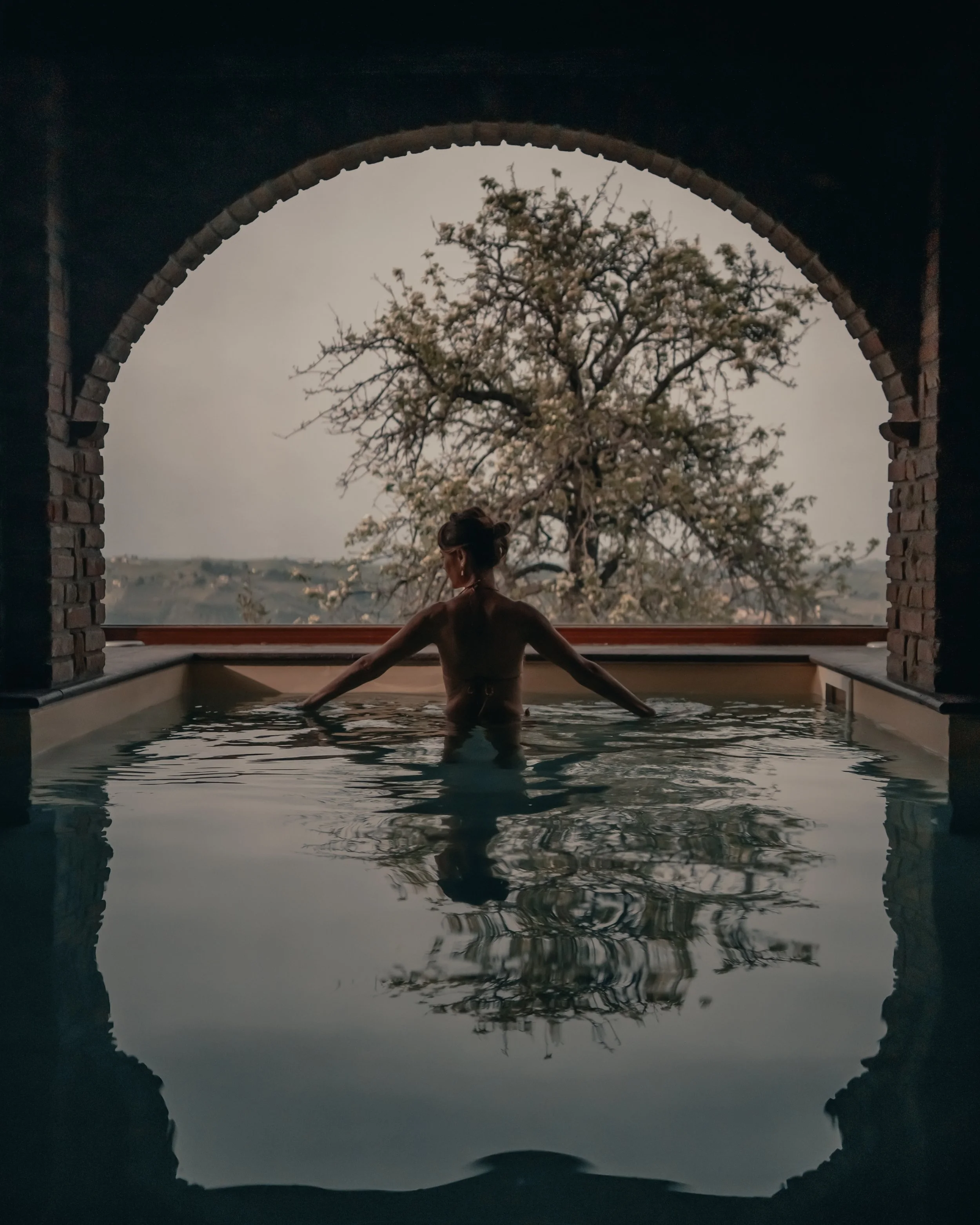 Donna in piedi nell'acqua di una piscina, vista attraverso un arco di pietra, con un albero fiorito sullo sfondo.