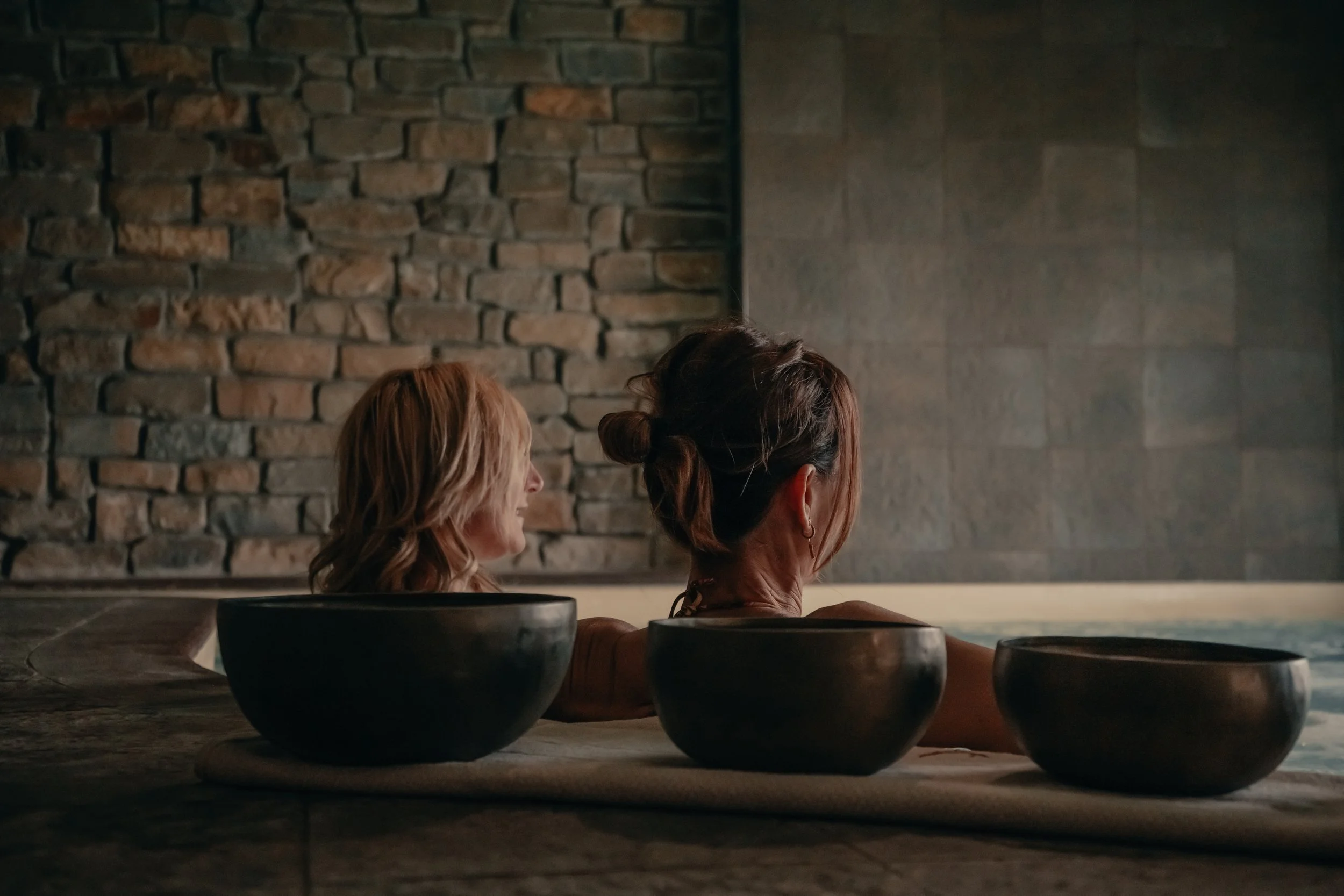 Due donne adulte immerse in una vasca termale o piscina, con un muro di pietra sullo sfondo, accanto a tre ciotole nere appoggiate su un asciugamano.