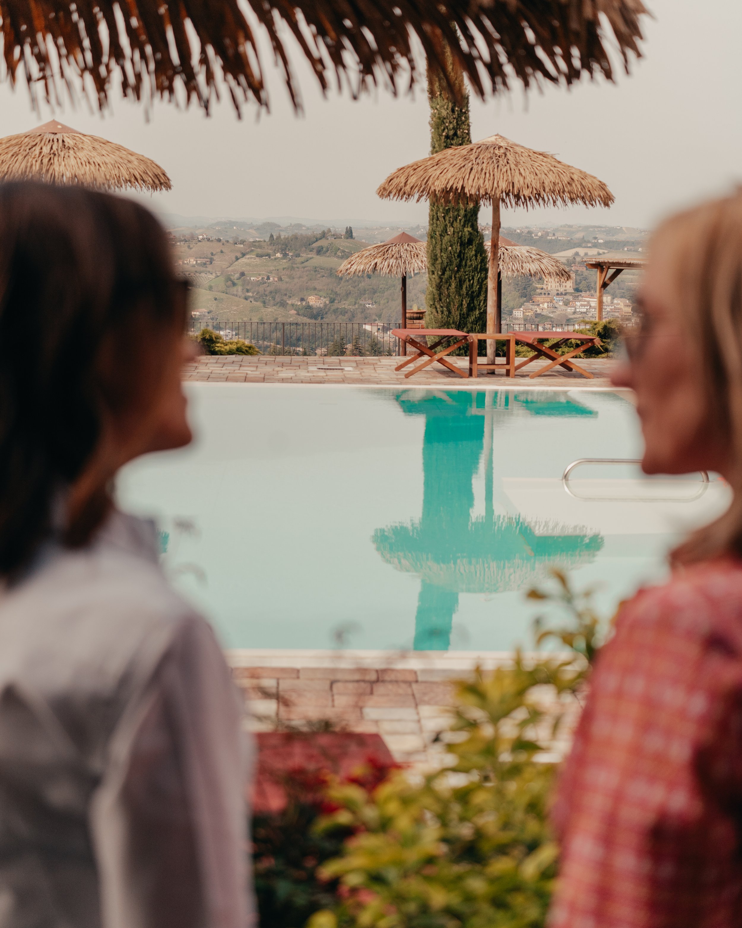 La foto mostra una piscina in trasparente e rilassante con uno sfondo di colline e case in lontananza. Sulla terrazza ci sono ombrelloni di paglia e sdraio.