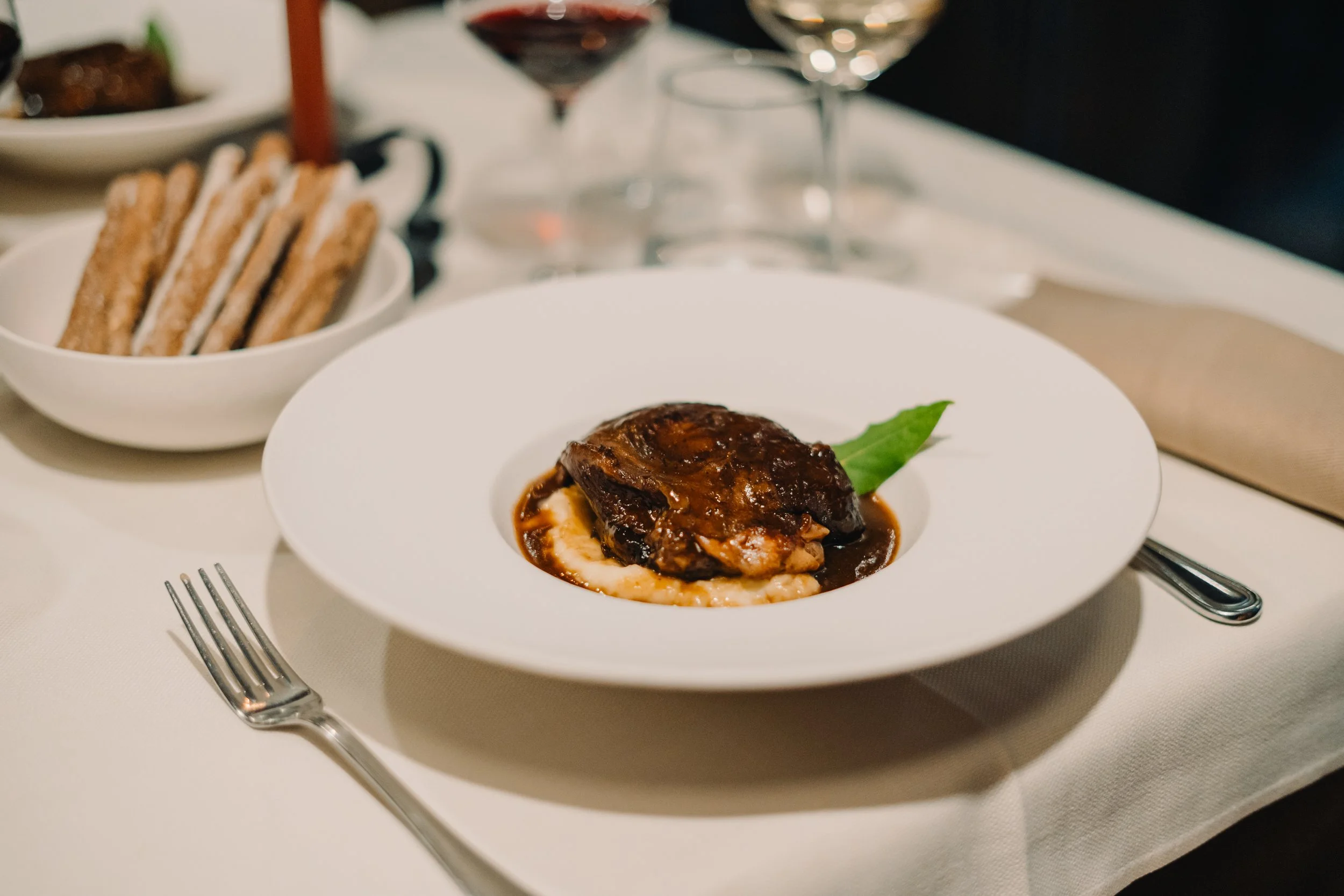 Piatti di cibo con un piatto principale di carne con condimento scuro e due bicchieri di vino rosso sul tavolo. Accanto, ci sono delle fette di pane croccante in una ciotola.