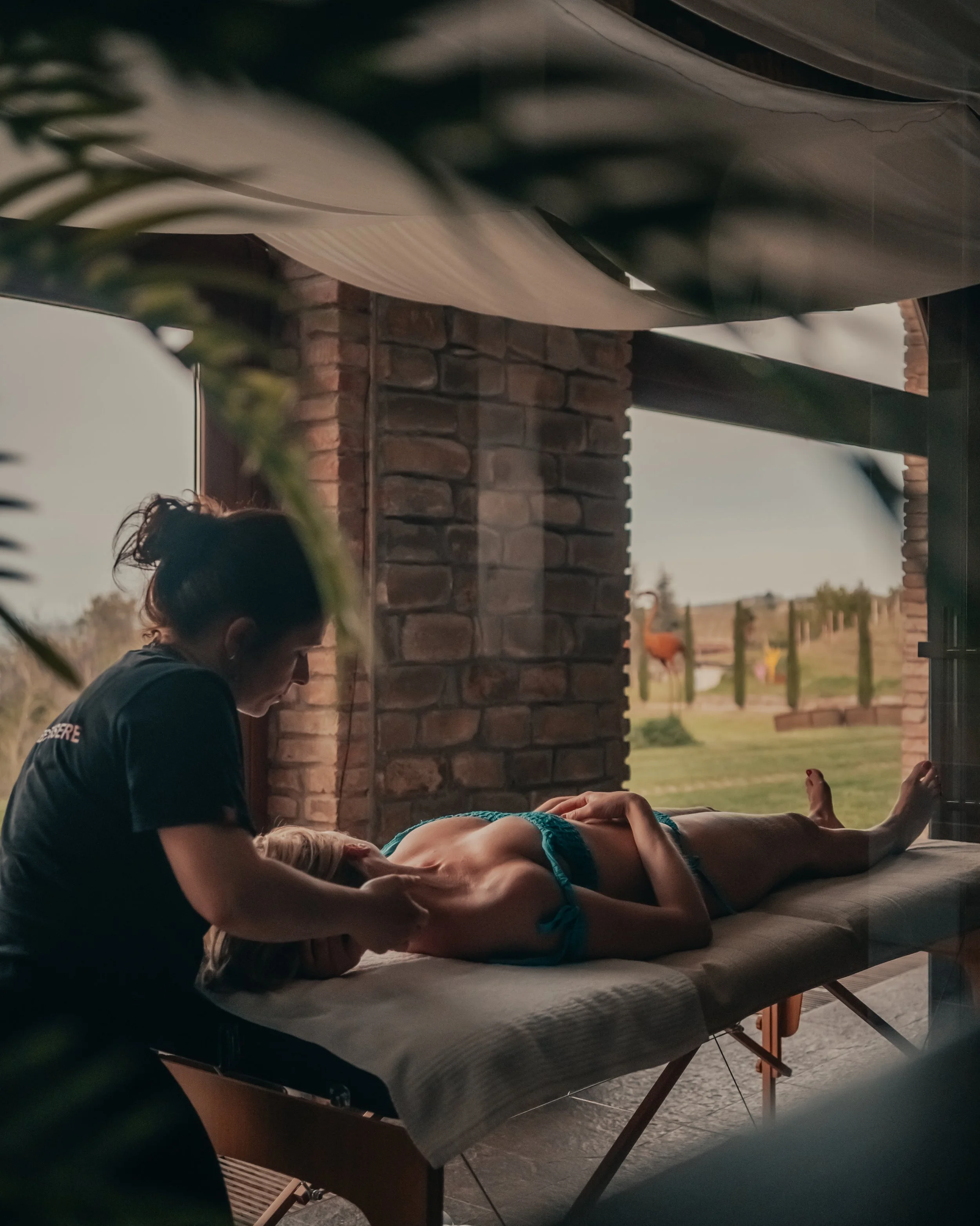 Massaggiatore che sostiene una donna sdraiata su un tavolo da massaggi, in un ambiente con pareti di mattoni e panorama naturale visibile attraverso una grande finestra.