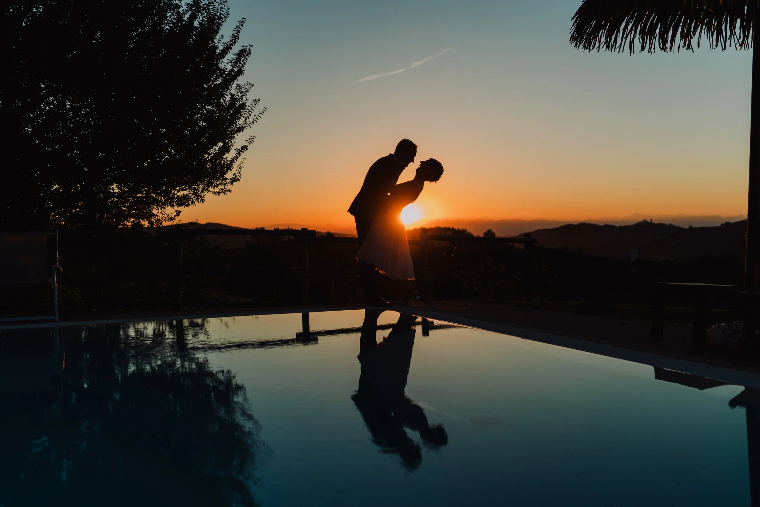 Silhouette di una coppia che si bacia al tramonto, con riflesso nell'acqua di una piscina, alberi e il cielo variopinto sullo sfondo.