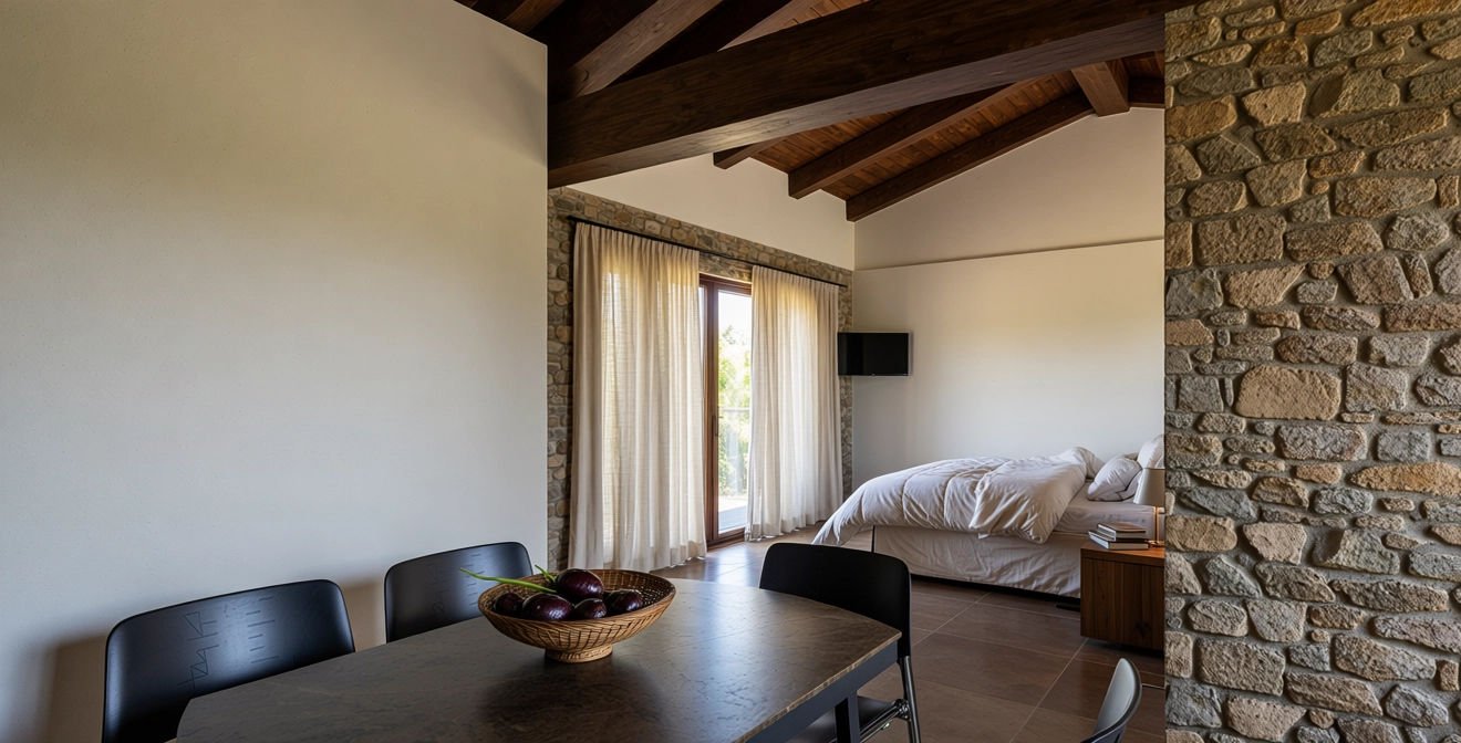 Interno di una stanza con soffitto a travi in legno, pareti in pietra e una finestra con tende chiare. In primo piano un tavolo con una ciotola di cipolle, sullo sfondo un letto con biancheria bianca e un piccolo televisore