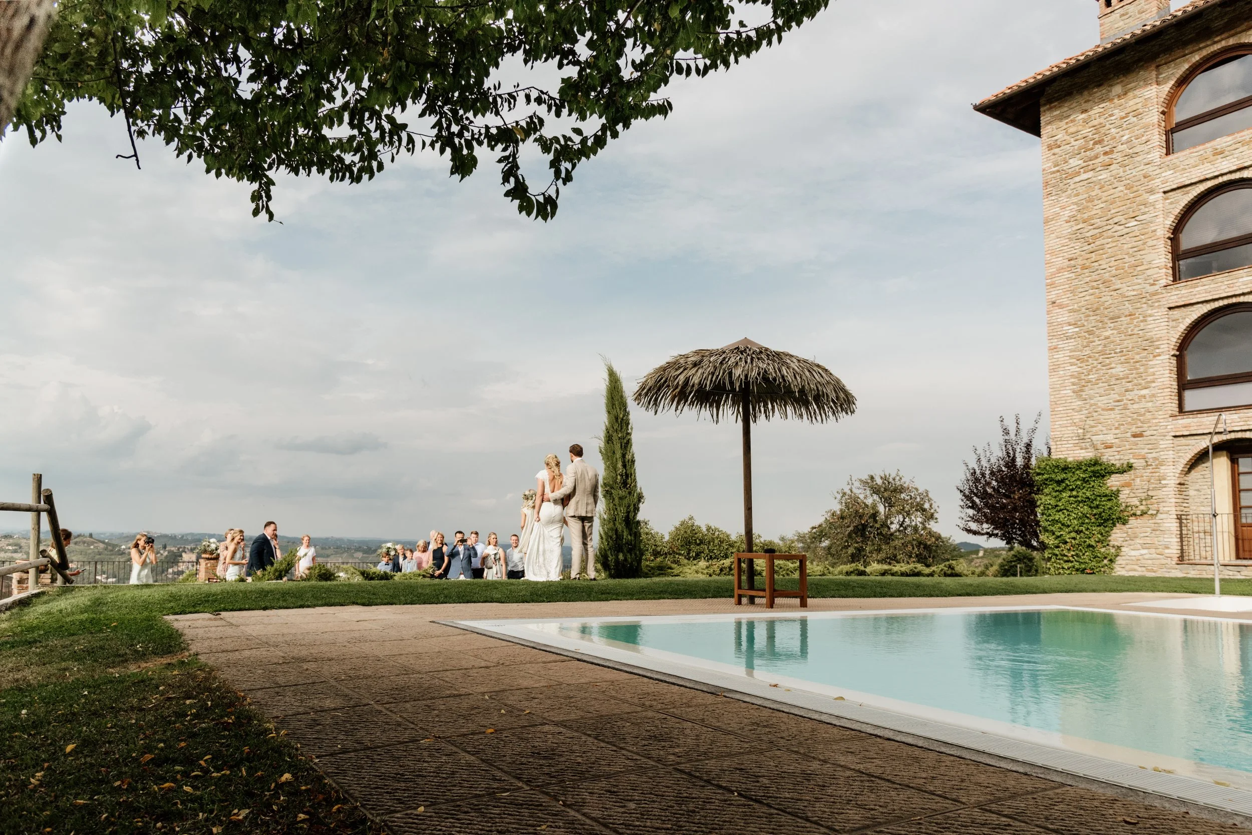 Gli sposi si tengono per mano durante un matrimonio all'aperto con molti invitati, vicino a una piscina con uno sfondo di cielo nuvoloso e un edificio in mattoni rustico.