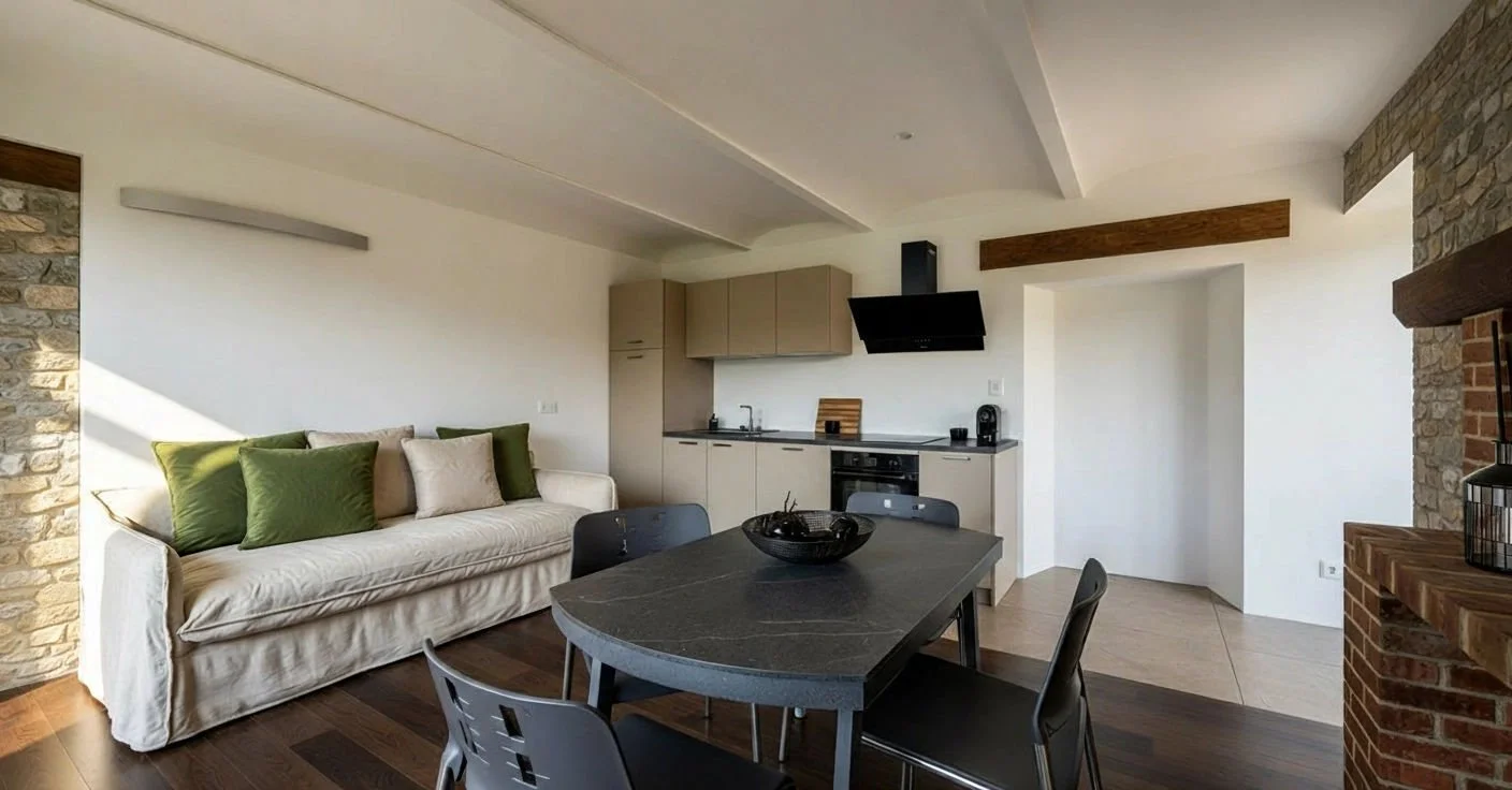 Soggiorno con divano bianco, cucina a vista con forno e cappa nera, tavolo con sedie, pareti di pietra e scaffale in legno.