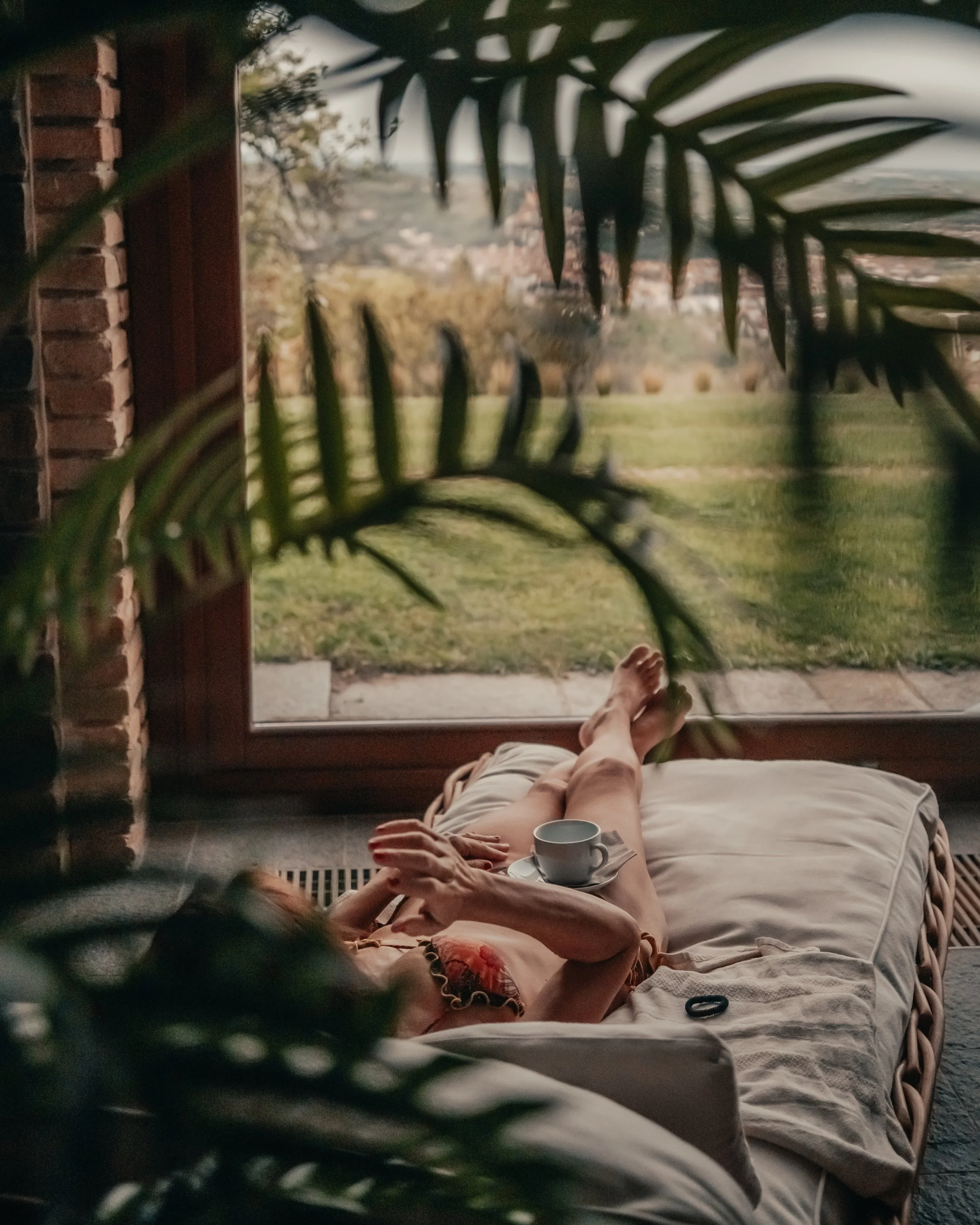 Una persona sdraiata su un letto con le gambe distese e un te' o caffè in mano, stesa di fronte a una finestra grande con vista su un cortile o un giardino all'aperto. La scena si svolge in un ambiente domestico, con un mazzo di foglie di una pianta in primo piano che copre parzialmente la visuale.