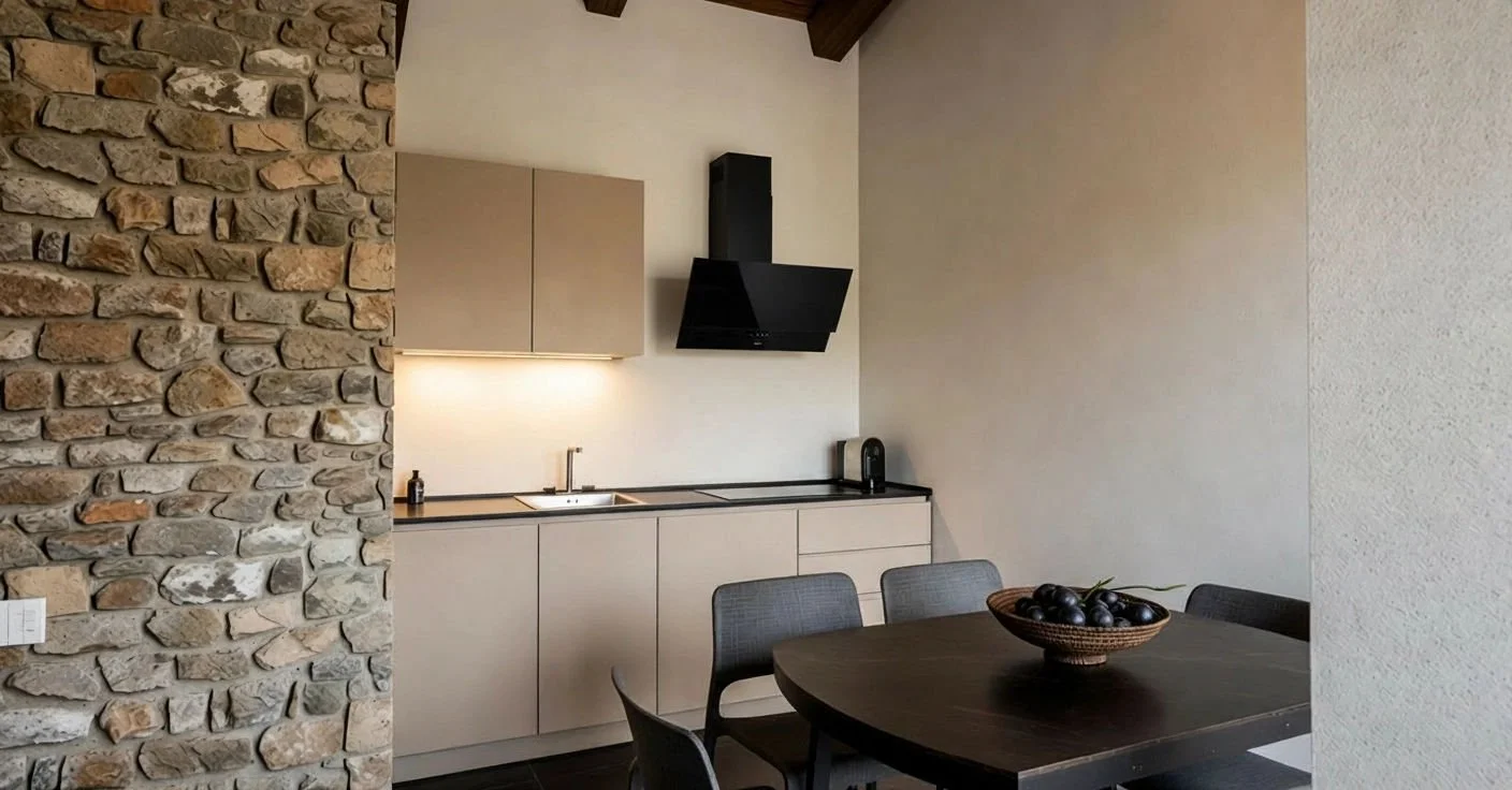 Cucina moderna con parete in pietra, top color beige, credenze in legno, piano cottura nero, cappa aspirante nera, tavolo da pranzo in legno scuro con ciotola di prugne