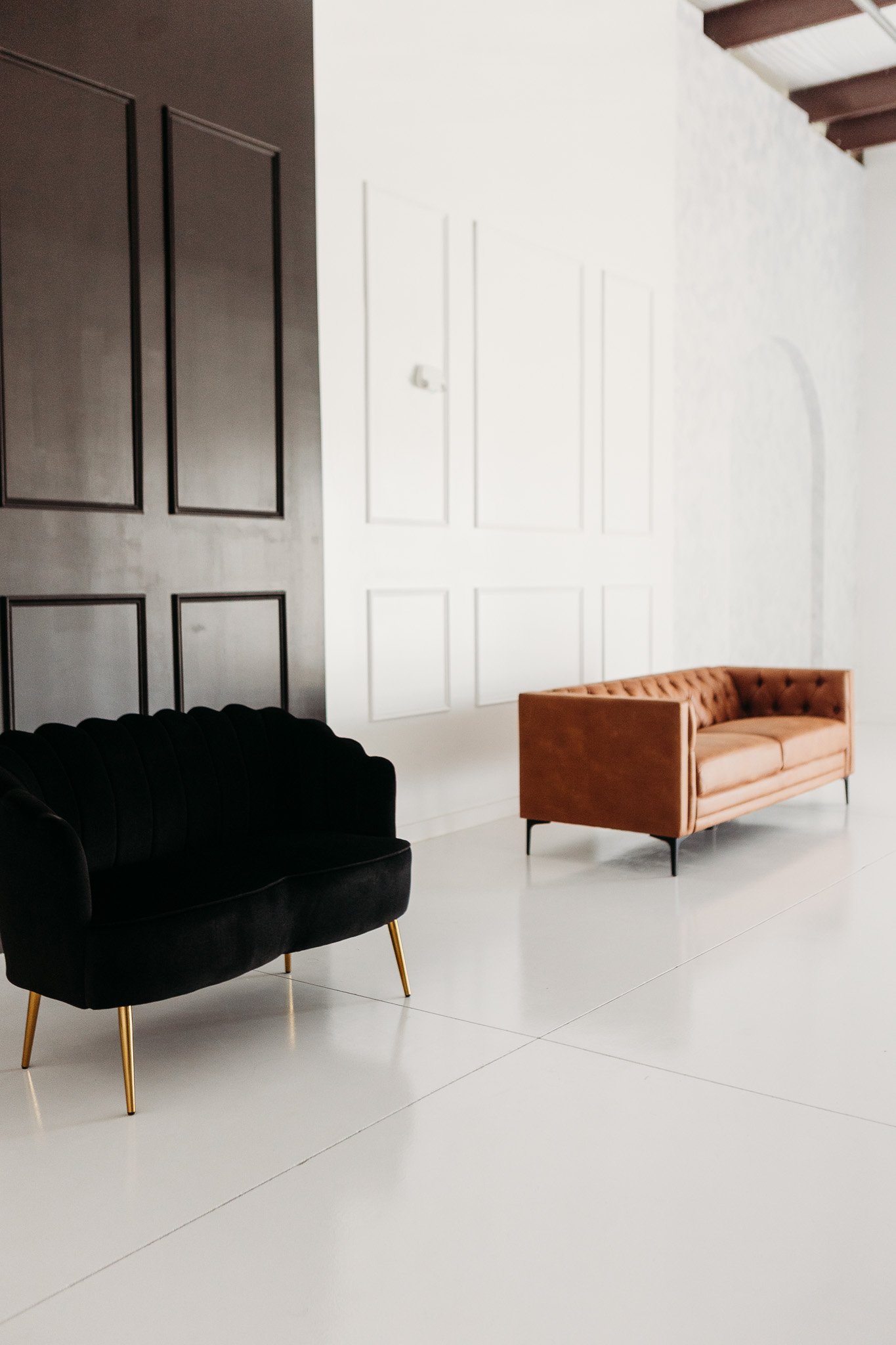 A living room with a black velvet armchair with gold legs, a pink velvet sofa, and a white wall with decorative paneling.