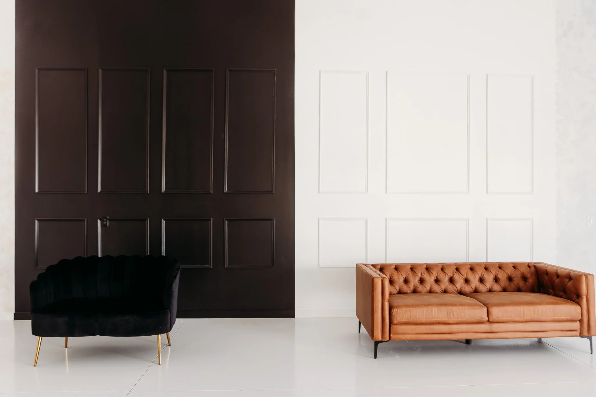 Interior of a room with two walls, one painted black and one white, featuring rectangular paneling. There is a black velvet armchair with gold legs on the left and a tan leather sofa on the right, both placed on a white tiled floor.