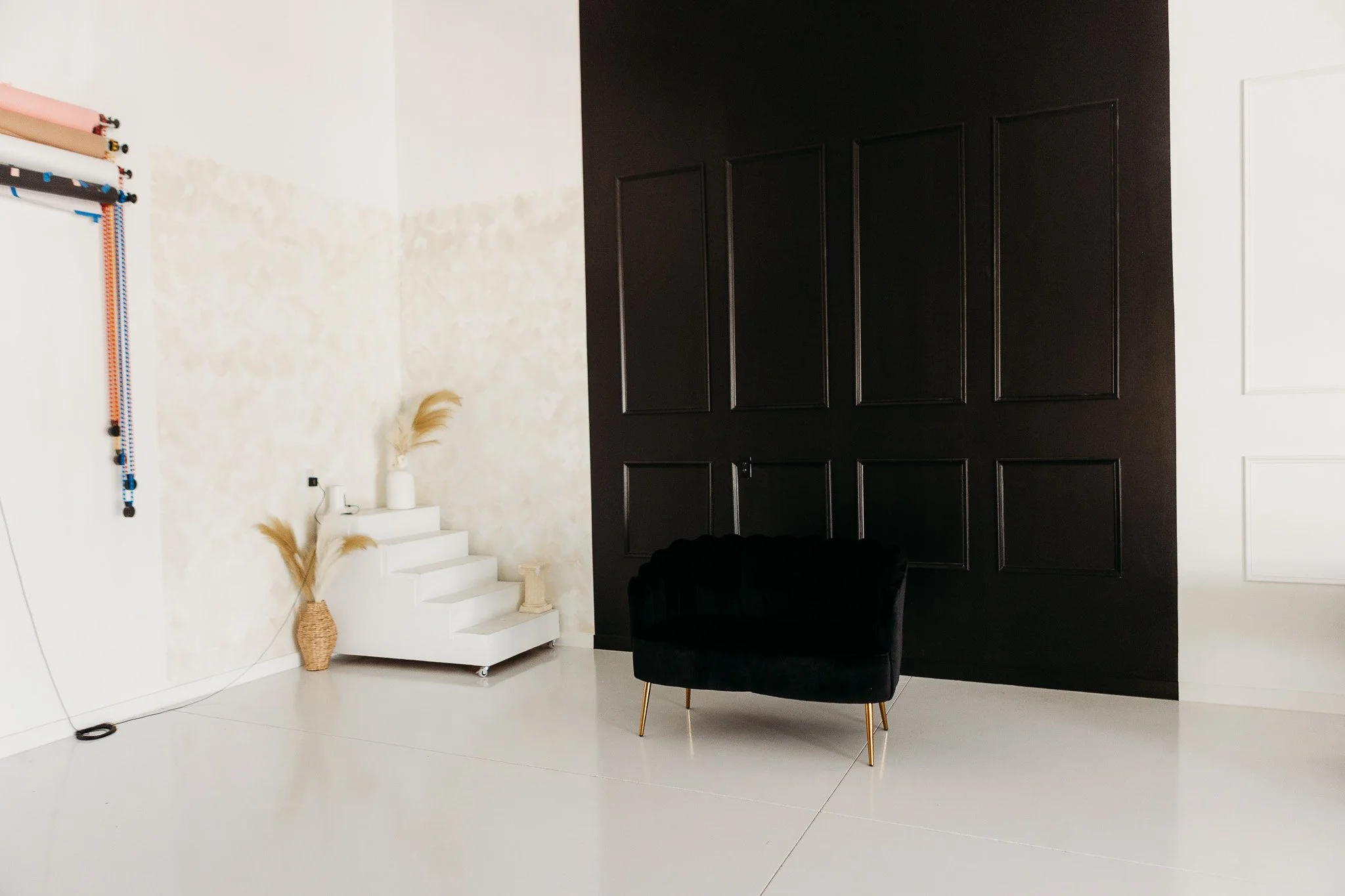 A modern interior with a black paneled wall, a white staircase, and a black chair with gold legs. Decor includes vases with dried plants and colorful hanging decor on the wall.