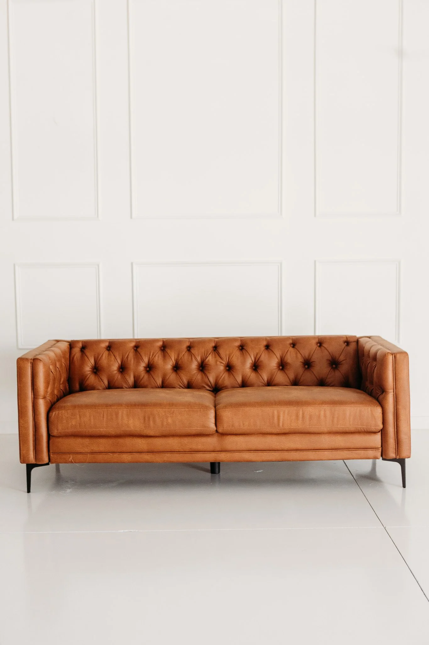 A tan leather tufted sofa with black metal legs against a white paneled wall.