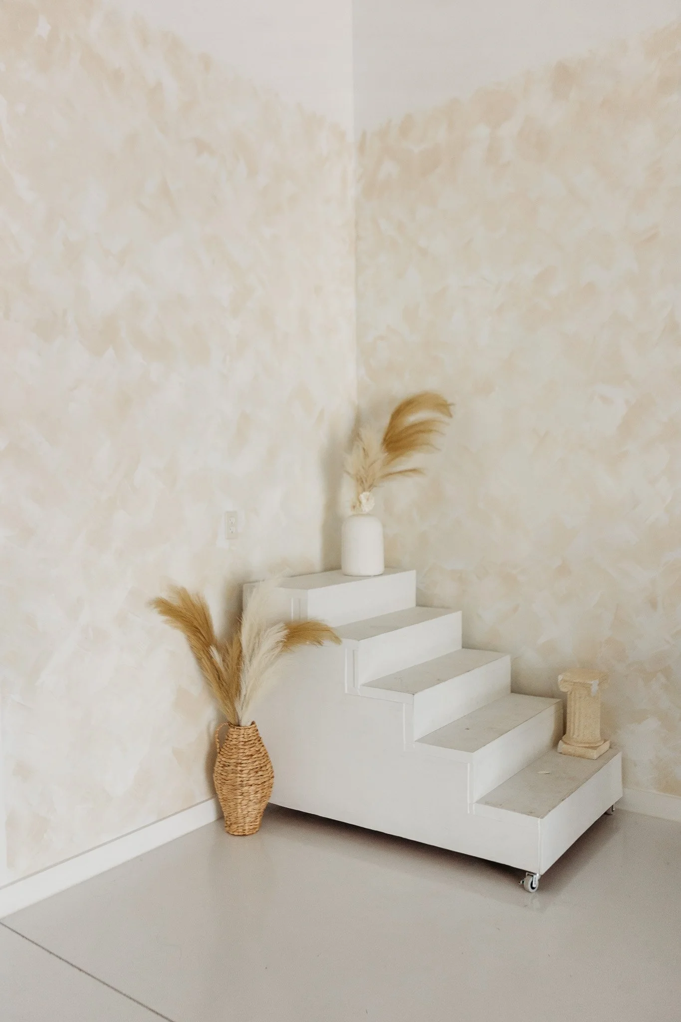 White staircase with rounded steps and caster wheels at the bottom, placed against a beige textured wall. A tall wicker vase with dried pampas grass is positioned on the left side of the staircase. There is a small, ornate beige sculpture on the right side on the floor.