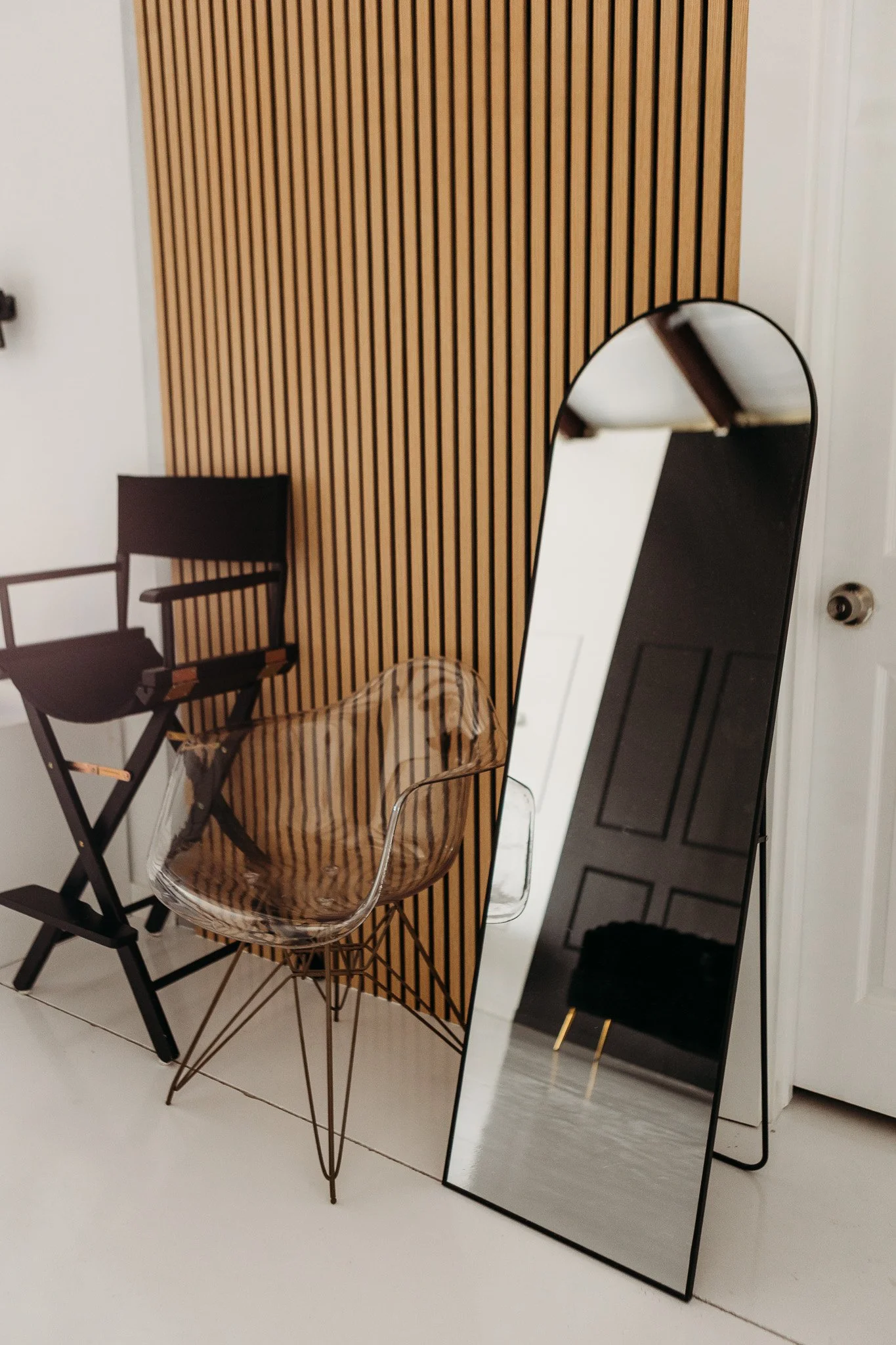 Interior decor with wooden slat wall, black chair, clear plastic chair, and a mirror reflecting a black door and white walls.