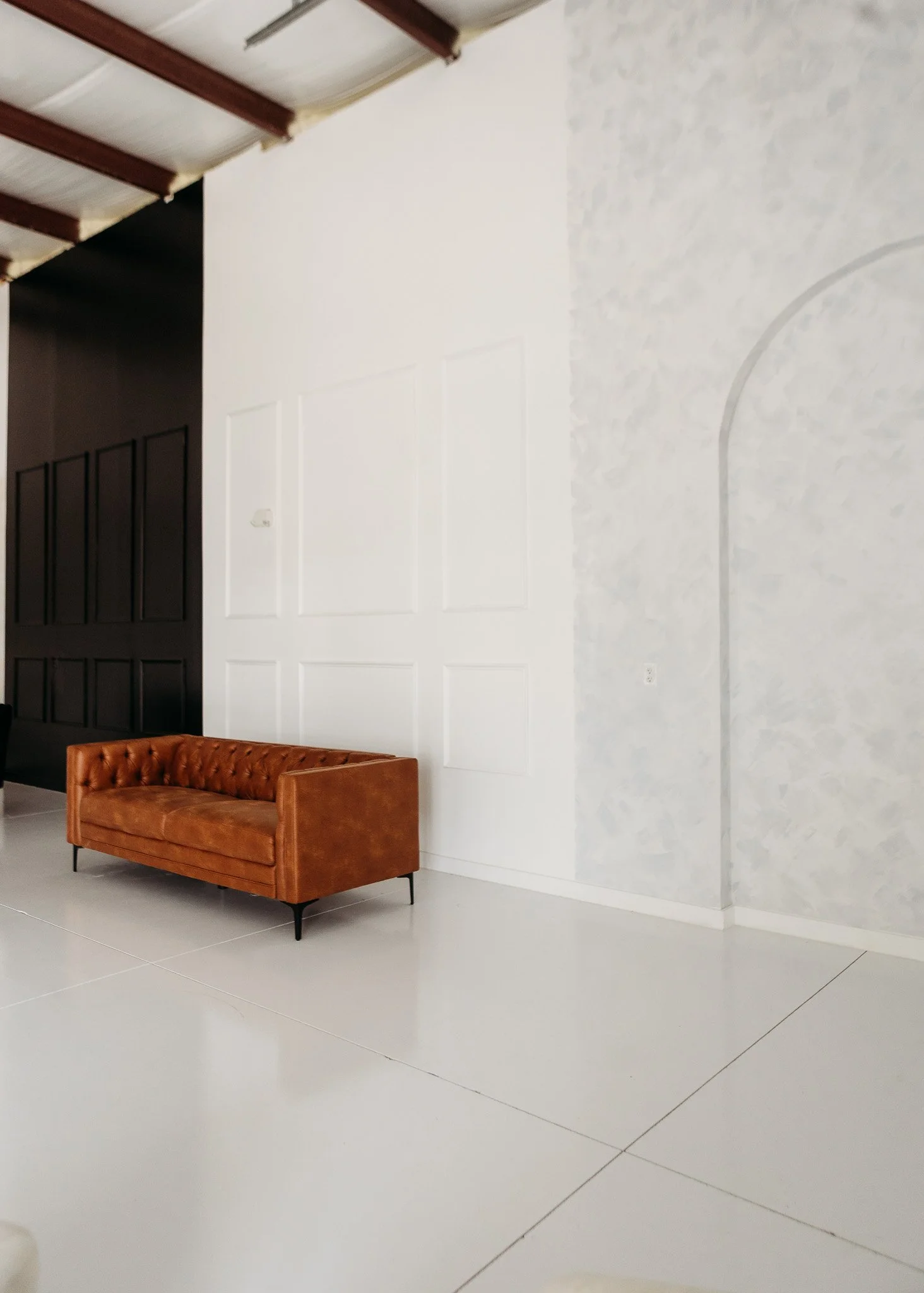 A brown leather sofa with button tufting sits on a white tiled floor at the bottom of the image. The background features a white wall with decorative rectangular paneling, and the room has a high ceiling with visible wooden beams. There is also a small white wall outlet near the floor.