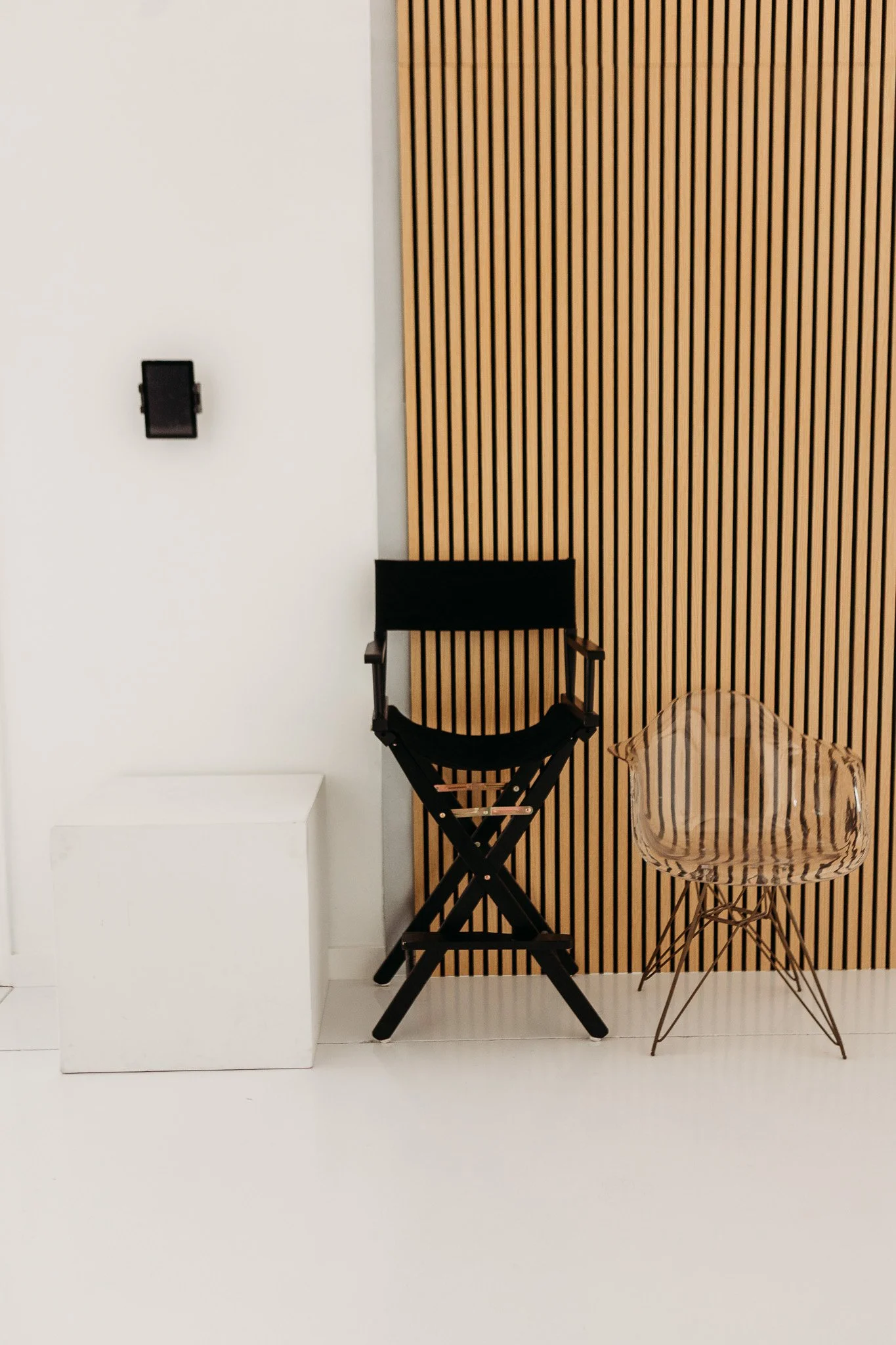 Interior scene with a wooden slatted wall, a black director's chair, a clear plastic chair, a white cube, and a white wall-mounted device.