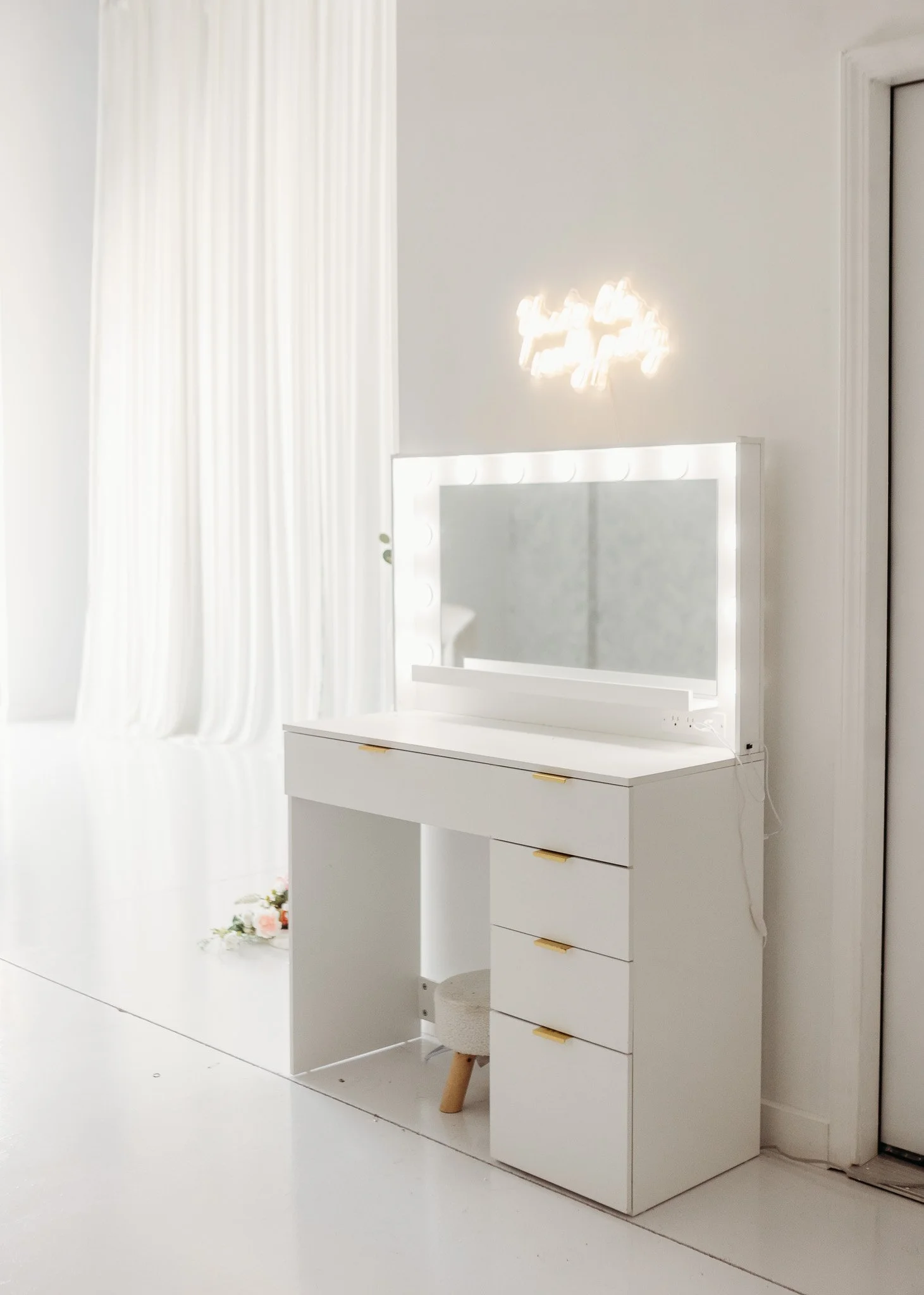 A white vanity with lit mirror and drawers, placed in a bright white room with curtains and flowers on the floor.