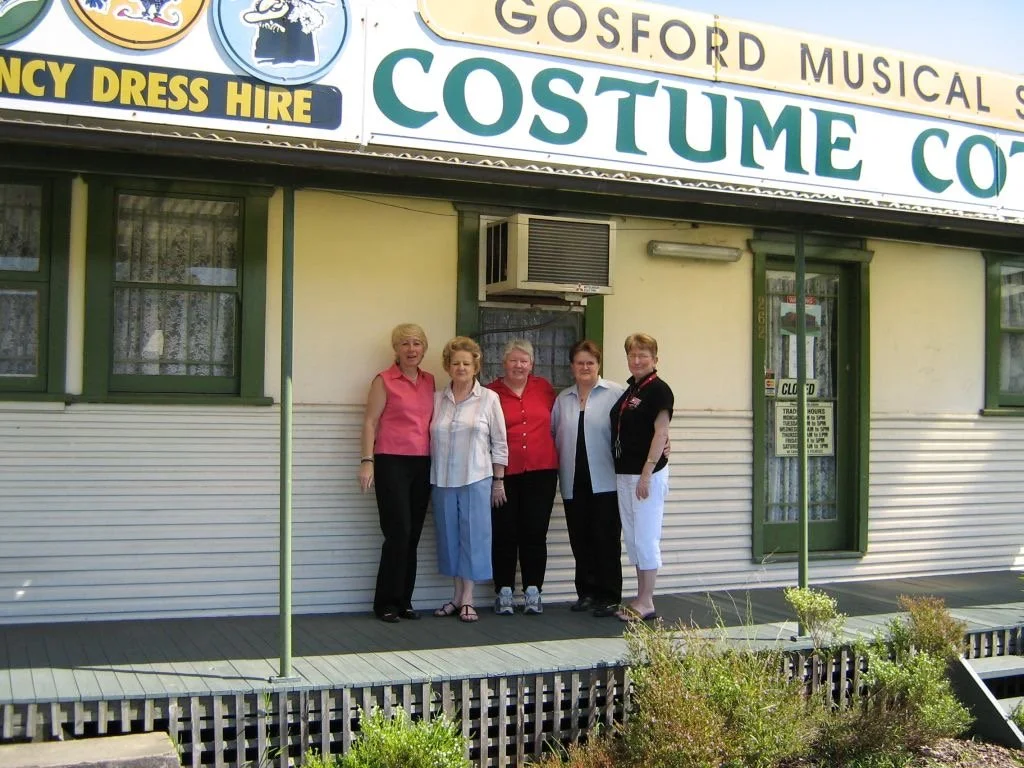 Four women standing in front of a costume rental store with a large sign that reads 'Gosford Musical' and 'Costume Co.' The shop has green window frames and a small porch with plants in the foreground.