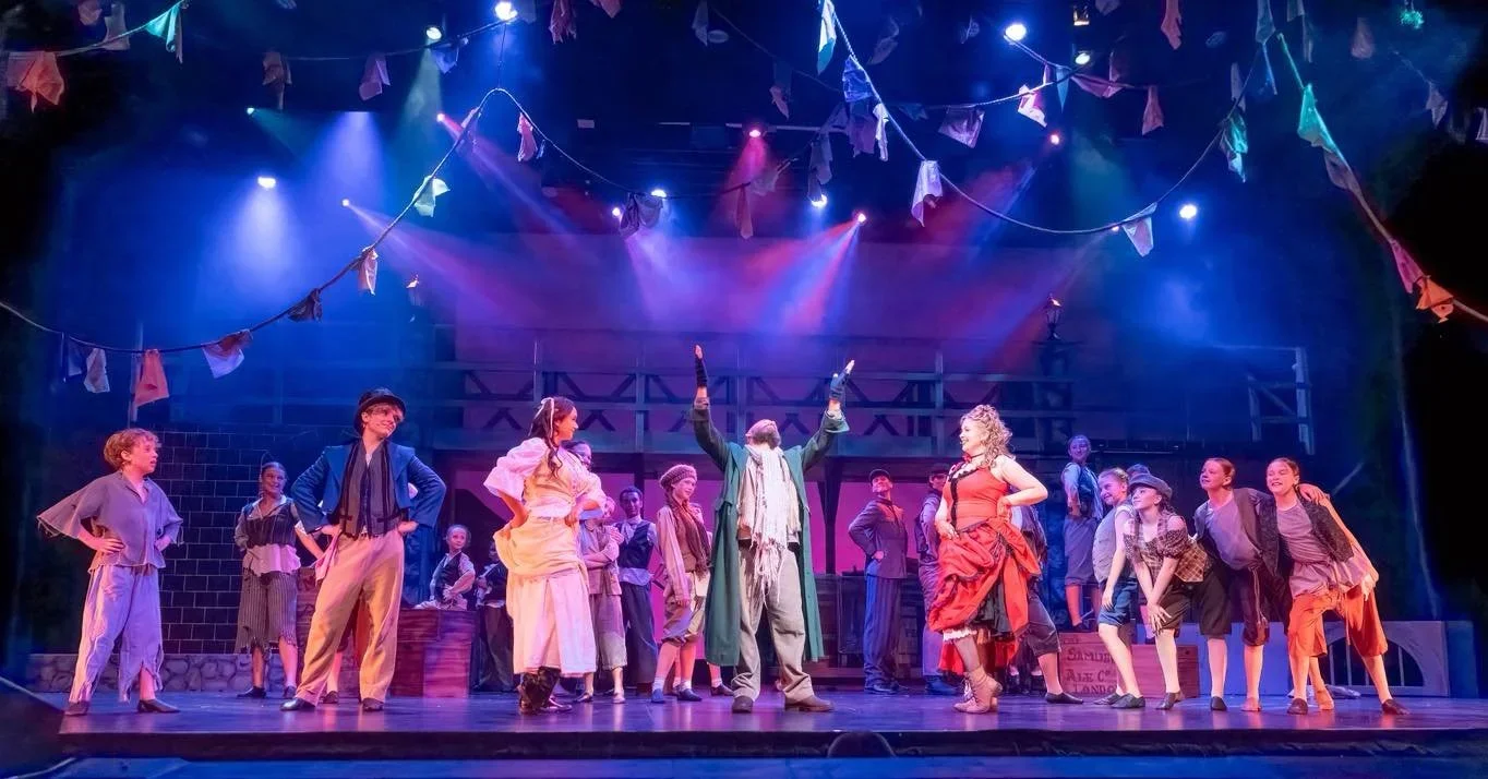 A theatrical stage scene with multiple actors dressed in period costumes, set against a vibrant, colorful backdrop with hanging banners, bright lights, and a rustic set design, portraying a lively story or musical.